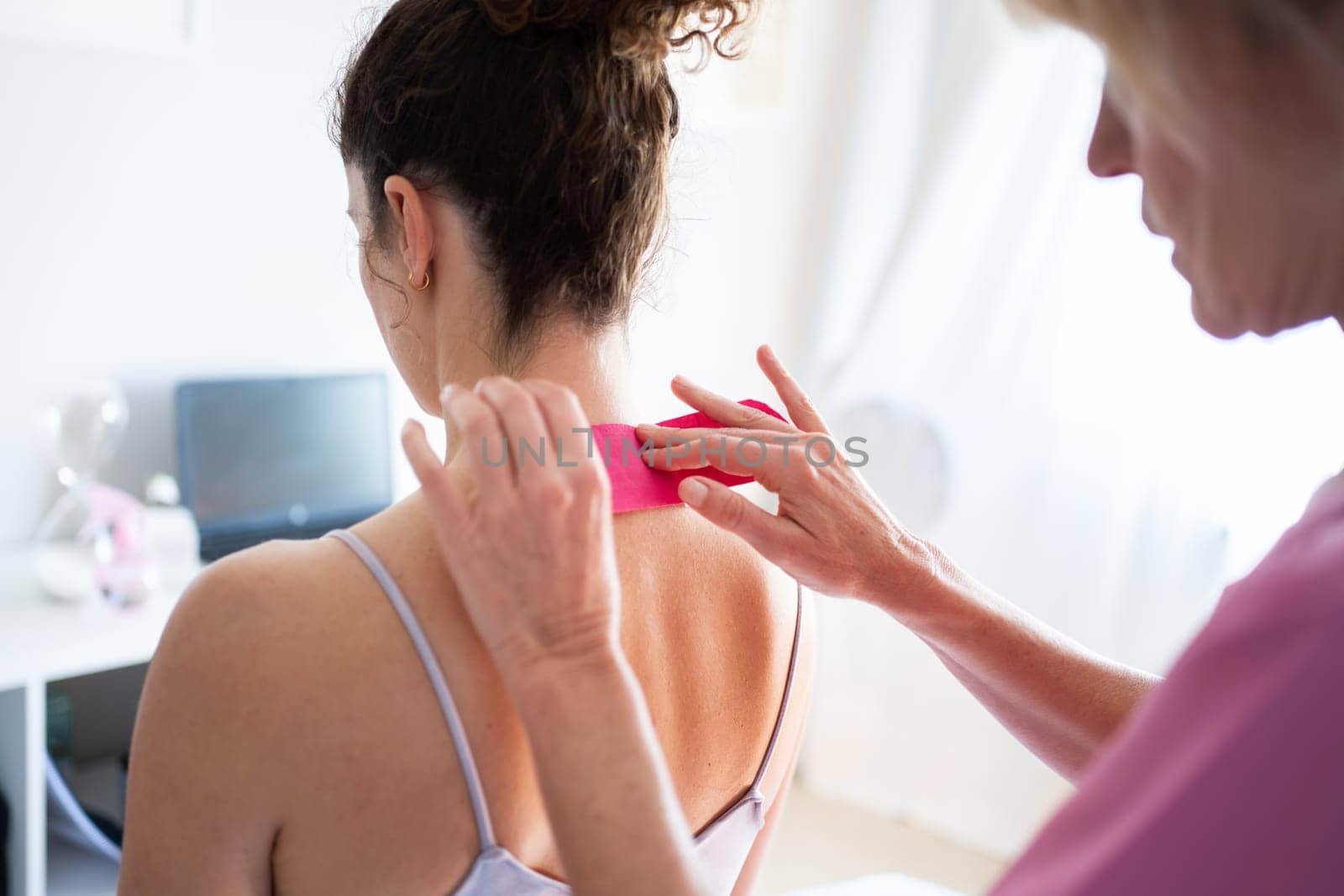 Crop professional osteopath applying kinesiology tape on neck of unrecognizable young female patient in inner wear during physiotherapy session in clinic