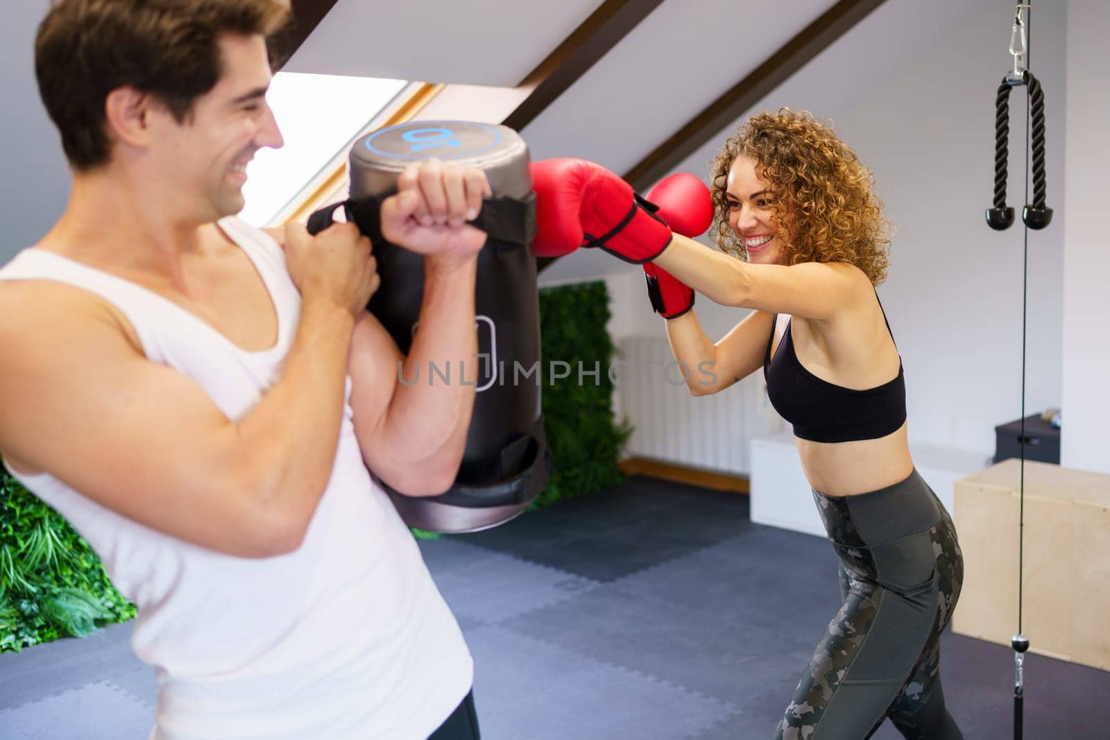 Young cheerful female fighter practicing punches while trainer holding heavy punching bag during boxing training in gym