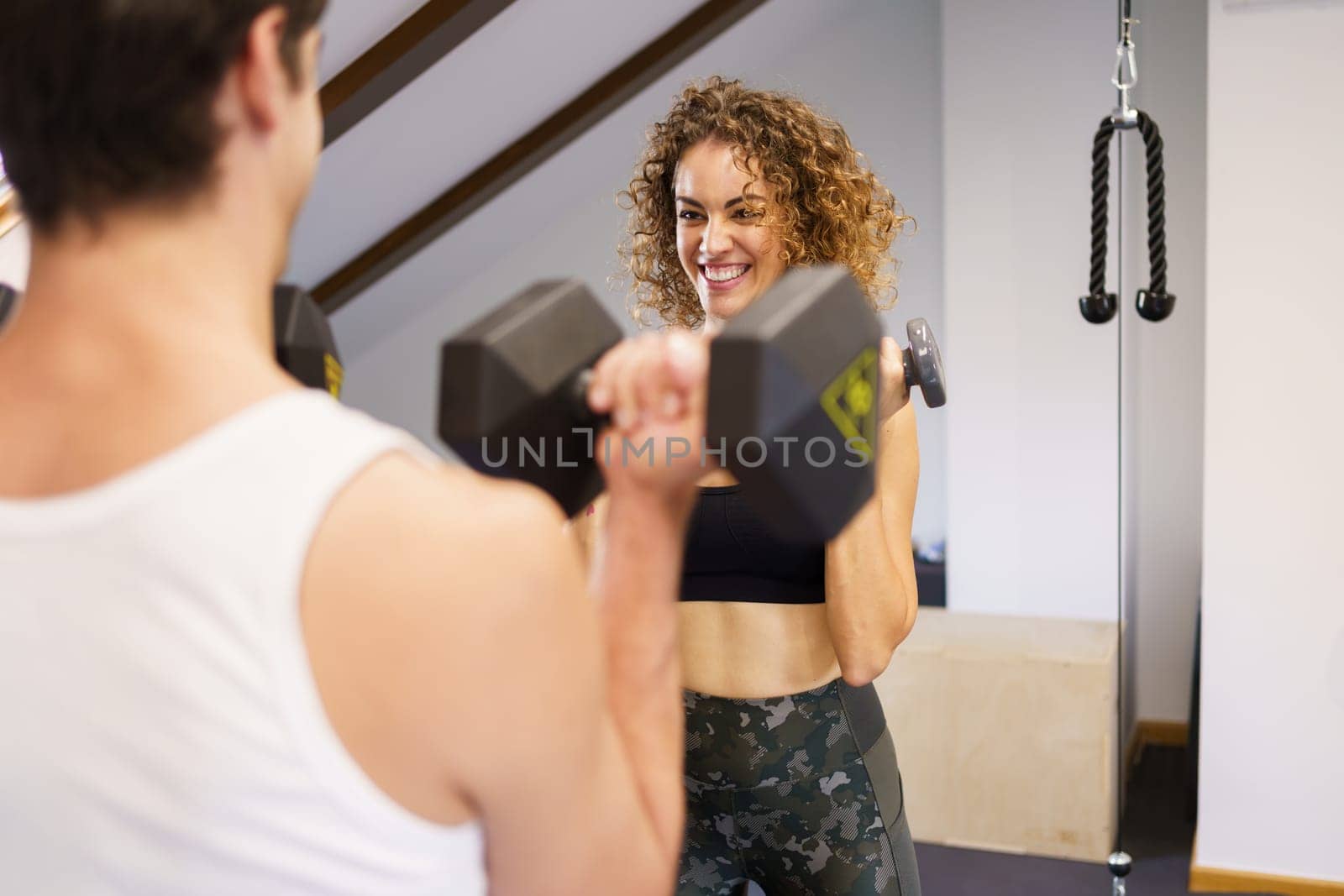 Young cheerful female athlete in tight sportswear lifting heavy dumbbells during workout with fitness buddy on weekend