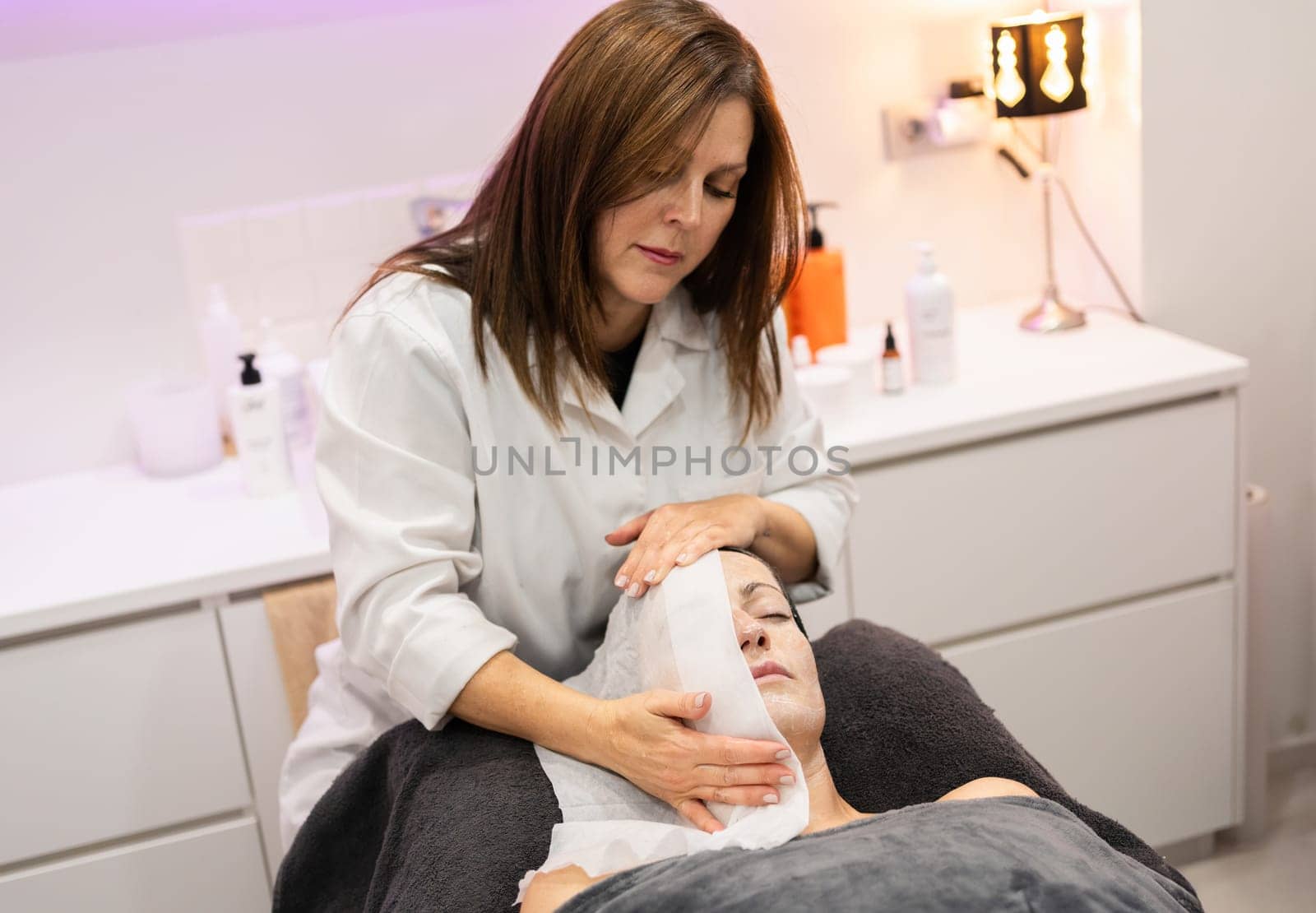 Professional female beautician looking down, while standing near beauty salon bed with anonymous client with closed eyes and treating face with hot water wet towel in lights