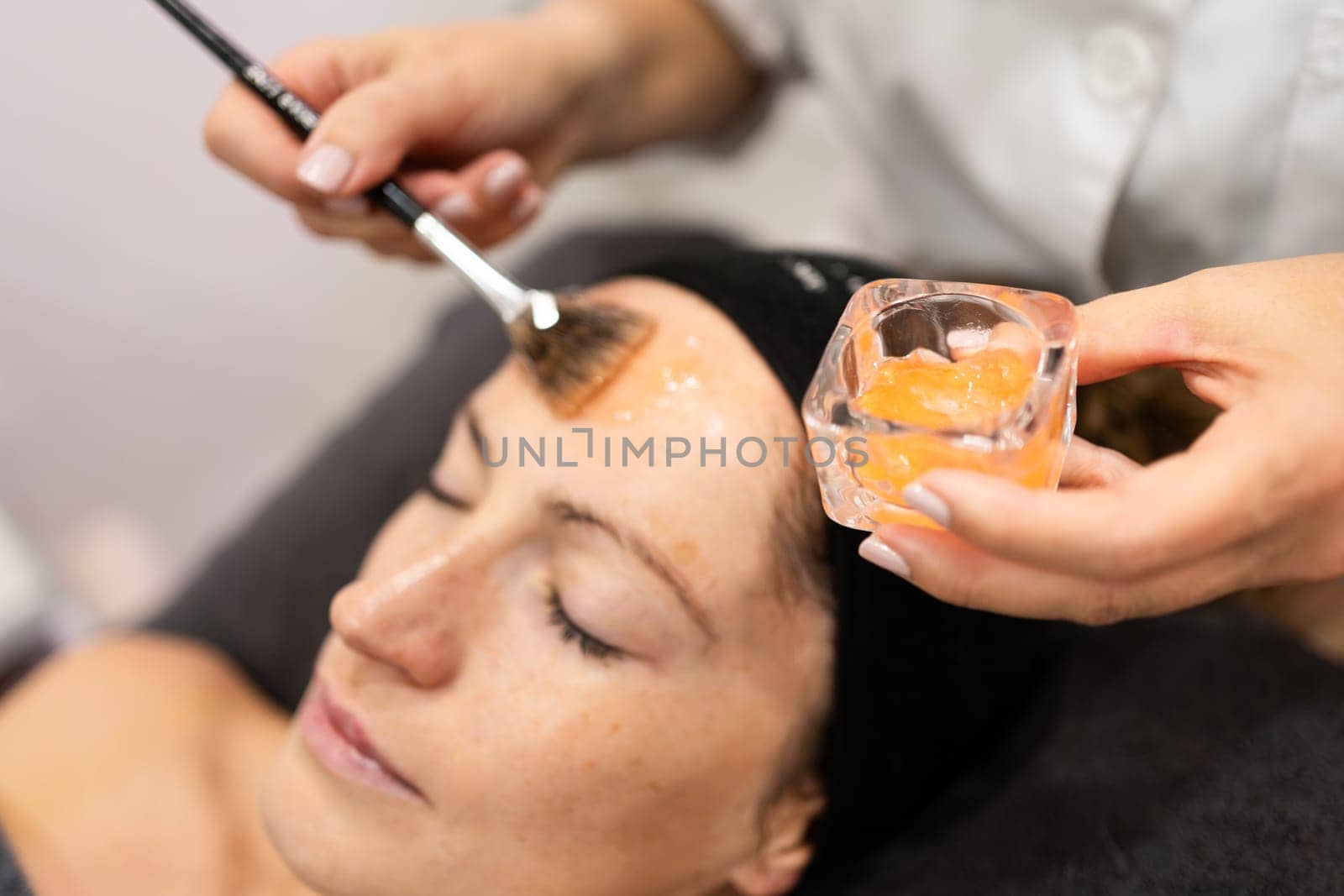 High angle of anonymous female beautician looking down, while standing near lady client lying with eyes closed on bed and applying moisturizing product with brush for skin treatment on forehead