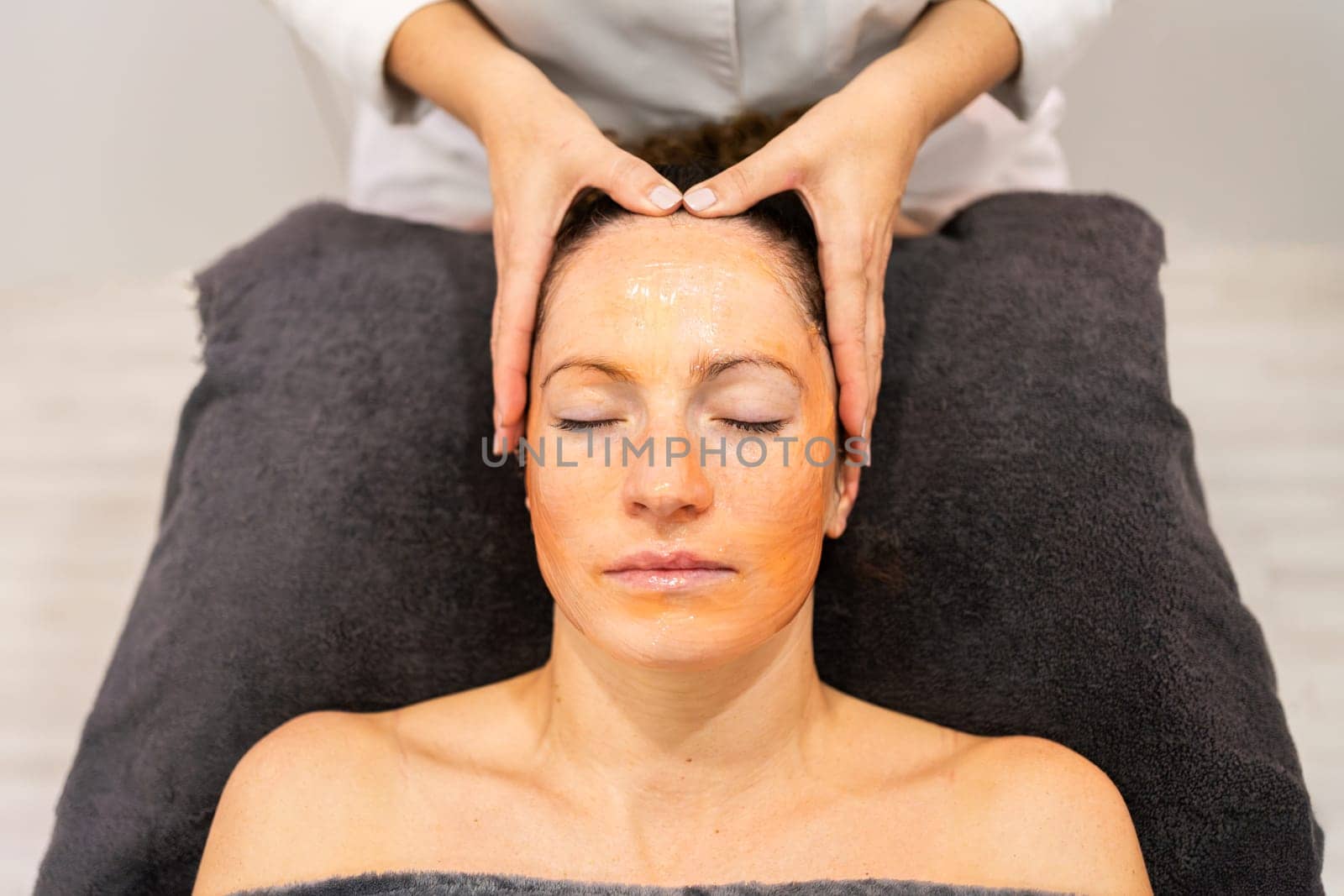 Crop cosmetologist doing facial massage of female customer, lying on towel with closed eyes and applying cleansing mask on face during skin care routine in beauty center