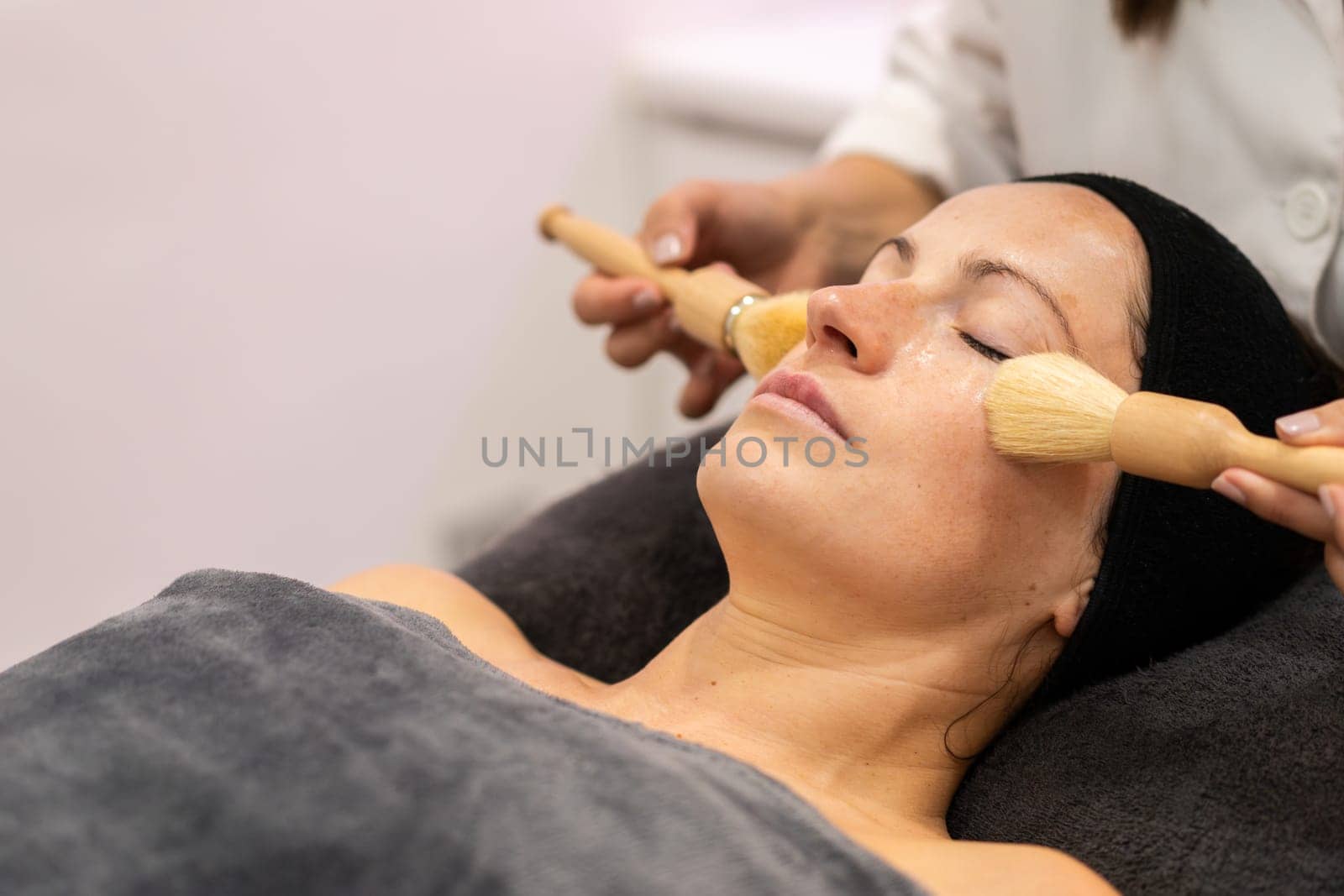 Crop beautician applying cosmetic cream on face of female customer with brushes during skin treatment in modern beauty center