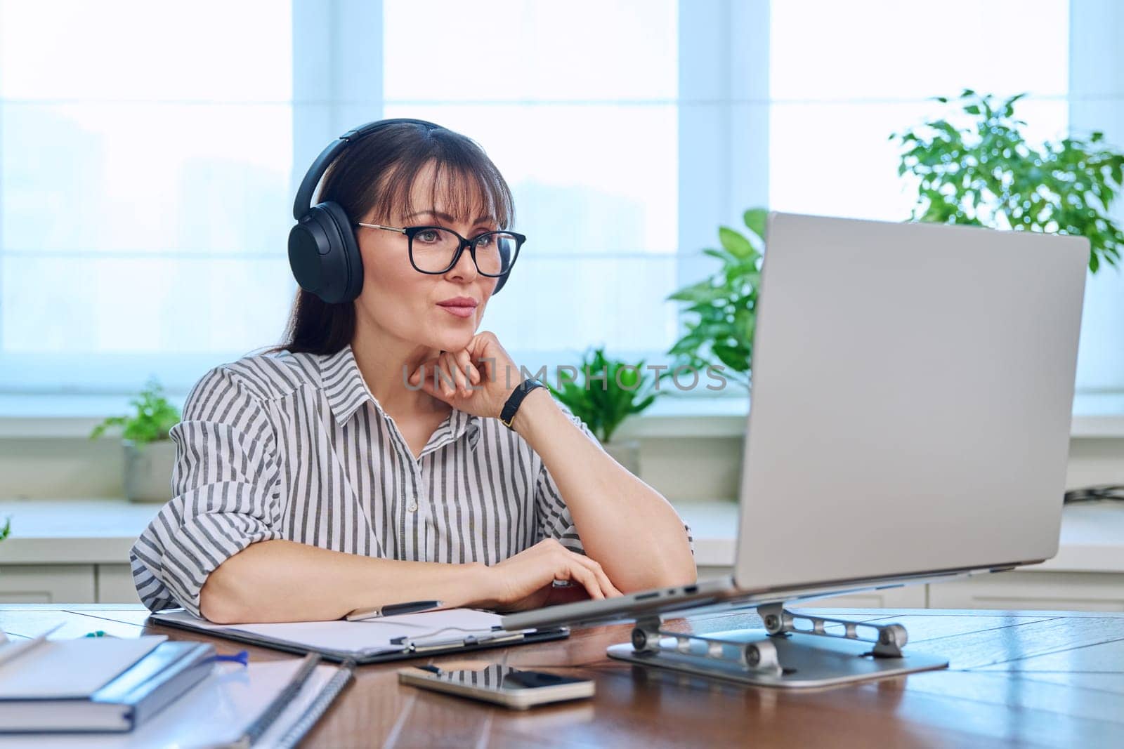 Middle-aged woman in headphones working at computer in home office by VH-studio