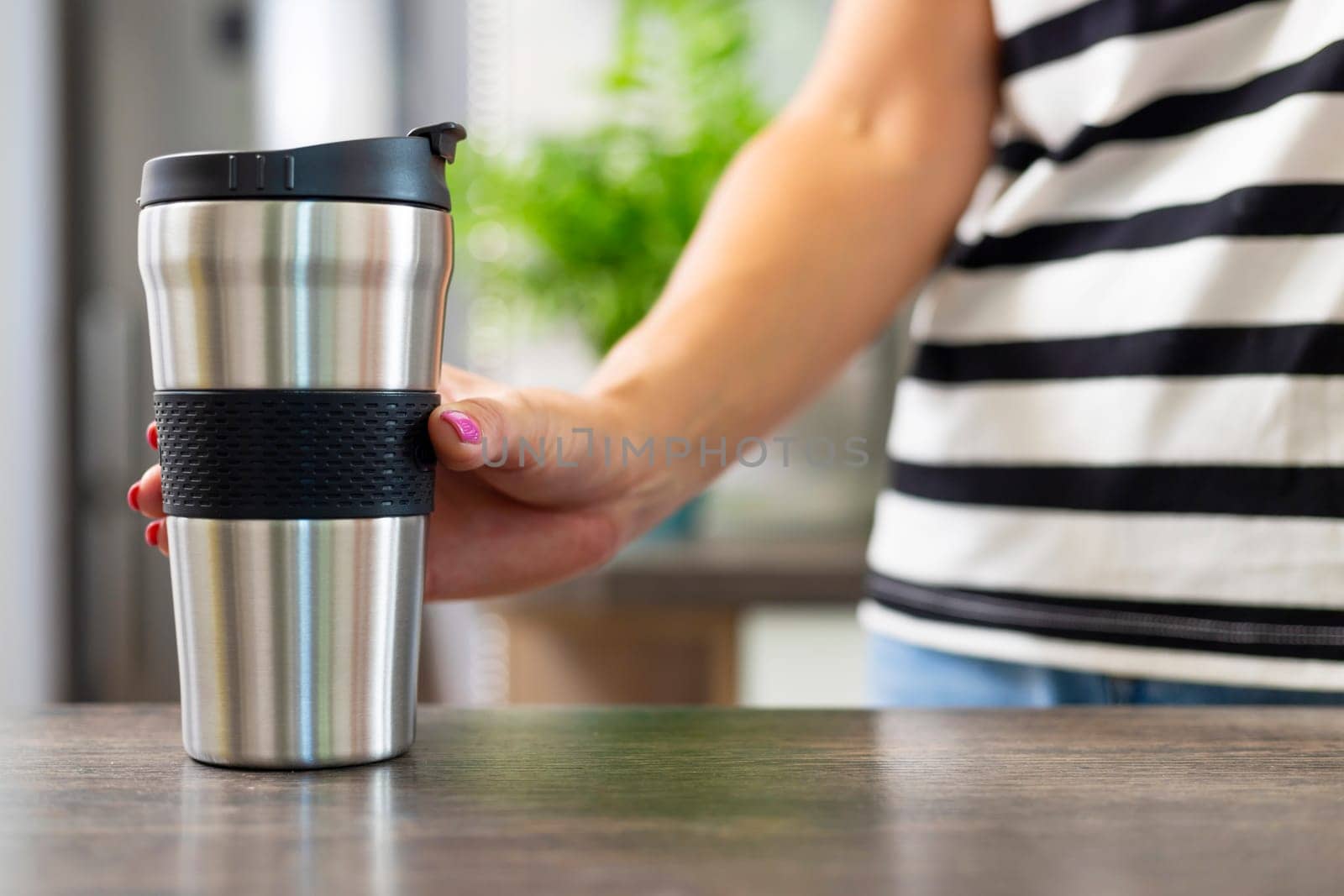 Close-up of a person's hand holding a stainless steel travel coffee mug with a black grip, wearing a casual striped shirt.