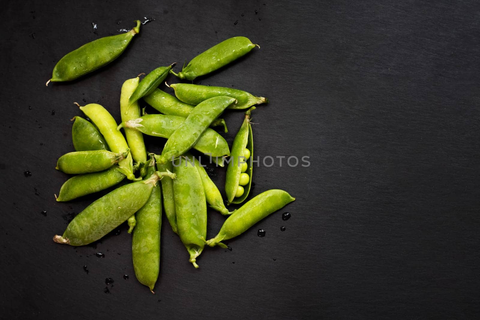 Fresh green pea pods scattered on a dark slate background, some opened showing peas inside.