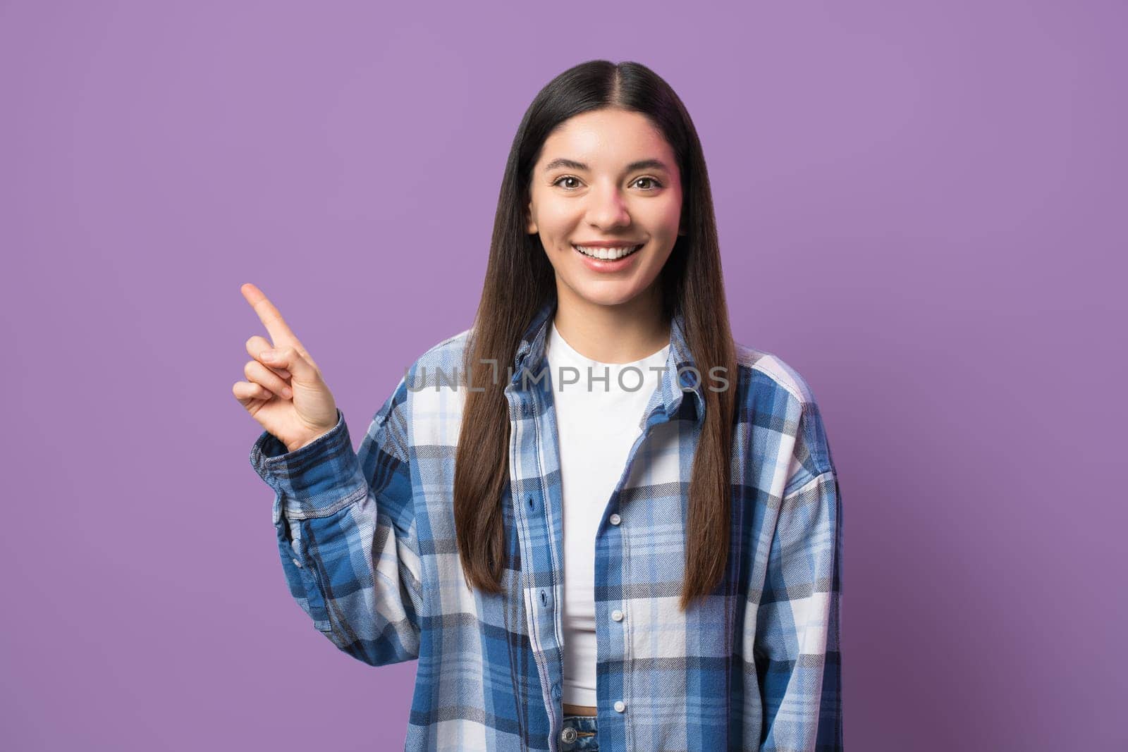 Multiethnic woman with attractive smile pointing with index finger up looking in camera against purple background.