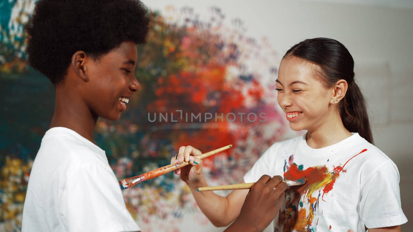 Smiling diverse children using paint brush painted color on each other white shirt shirt at colorful stained wall in art lesson. Represent exchanging experience, learning each other. Edification.