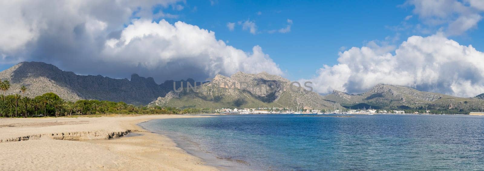 A breathtaking panoramic landscape where a lush palm oasis fringes a pristine beach leading to a clear sea, with majestic mountains looming under a dynamic sky