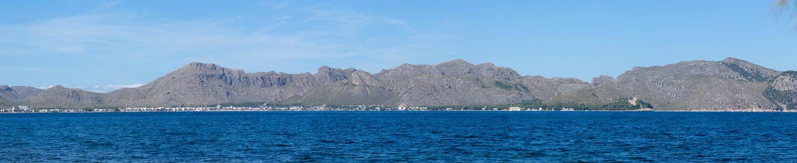 A panoramic view where the sapphire blue of the sea meets a dramatic mountain range, with a thin line of coastal habitations nestled at its base. The undulating peaks stretch across the horizon, under a clear sky, embodying the raw beauty of a natural seascape