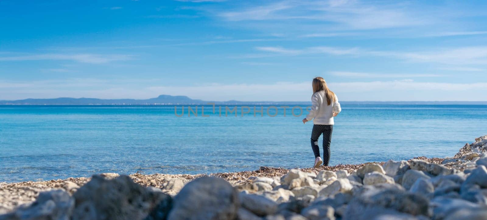 A child stands at the edge of a pebbled shore, gazing out towards the endless blue sea. The vastness of the ocean and the clear sky stretch out before them, inspiring wonder and the promise of discoveries beyond the horizon