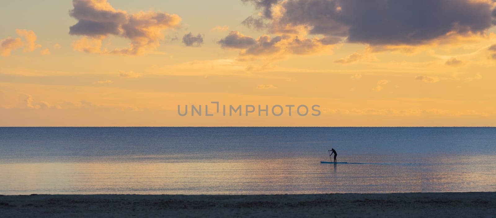 As the day winds down, a paddleboarder glides across the calm ocean, their silhouette a delicate contrast to the pastel hues of the sunset sky. This peaceful scene, set at the intersection of sea and sky, encapsulates the essence of tranquil seaside evenings.