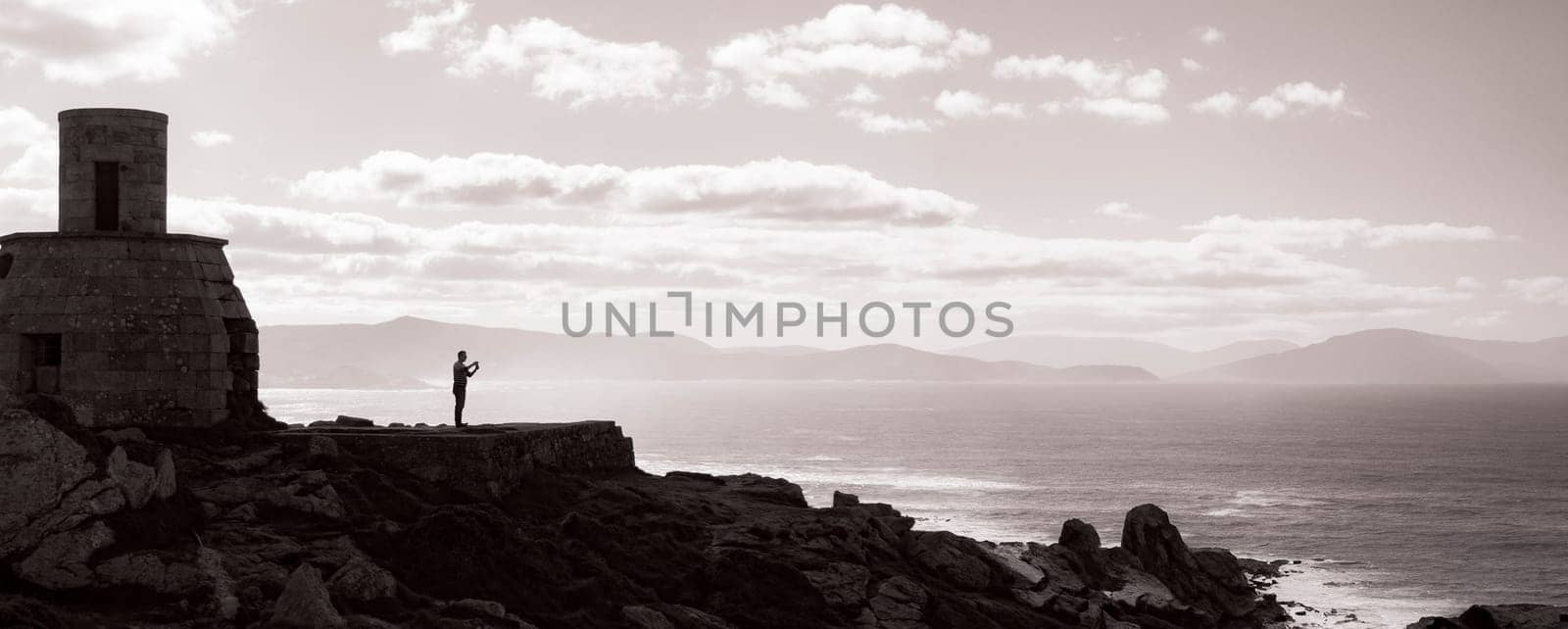 Captured in sepia tones, a solitary figure stands contemplatively at the edge of a rugged cliff, beside an old cylindrical watchtower. The expansive view of the sea and distant mountains adds to the scene's sense of isolation and introspective calm.