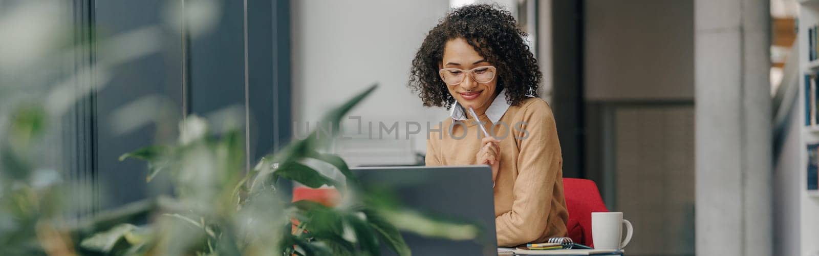 Stylish business woman in eyeglasses working on laptop while sitting in modern office near window