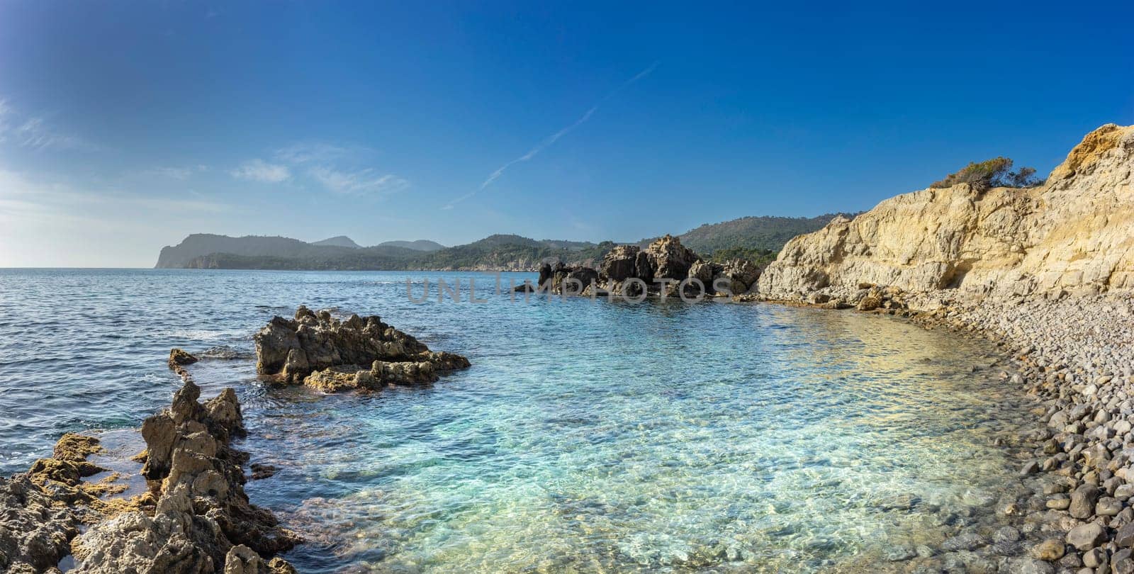 The rugged beauty of a rocky shoreline meets the clear, turquoise embrace of the sea under a wide azure sky. This panoramic view captures the serene meeting of land and water, highlighting the pristine clarity of the Mediterranean coast