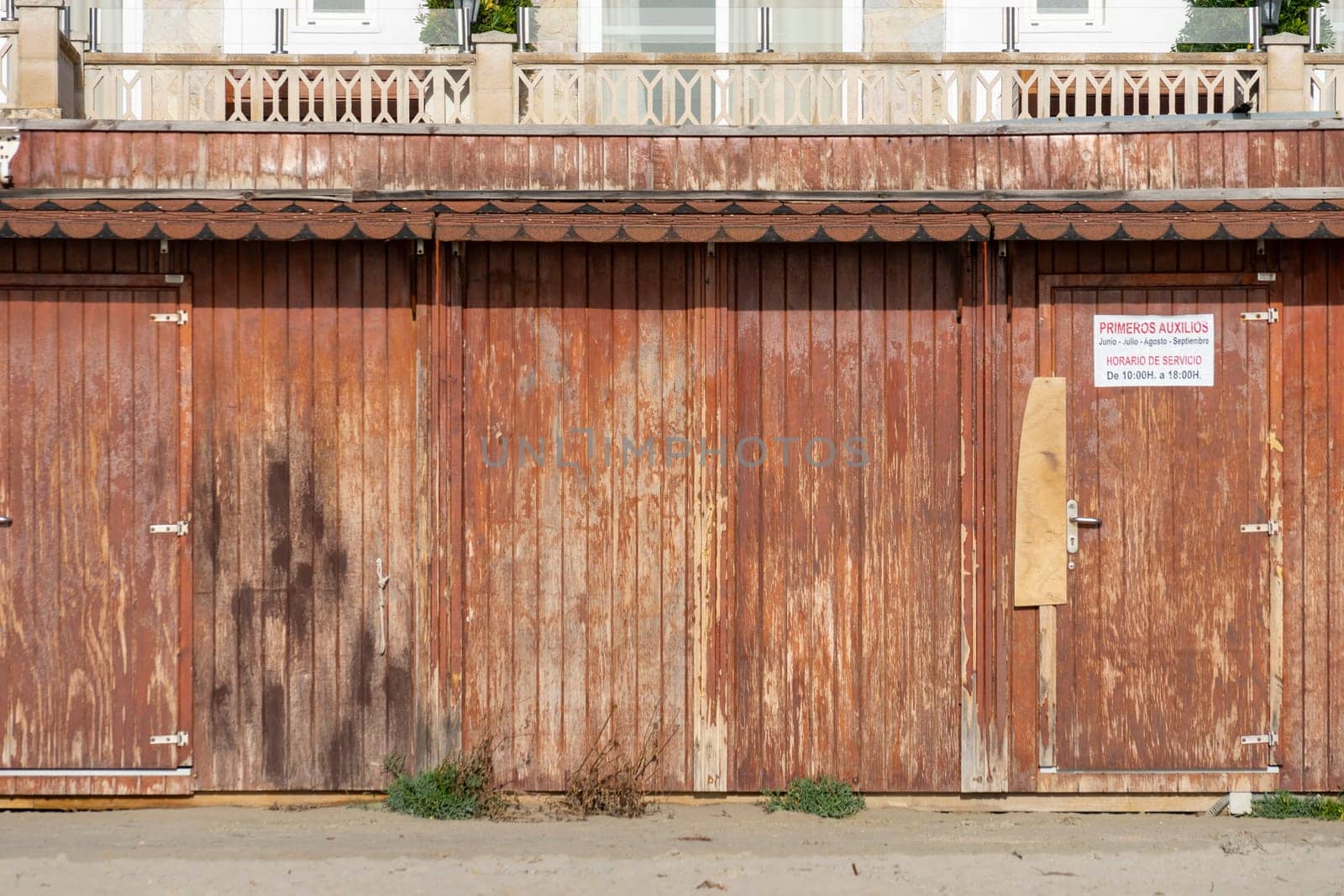 A row of rustic, weathered beach huts stands with a quiet history, their reddish-brown doors showing signs of salt and sun, juxtaposed against an elegant balcony above