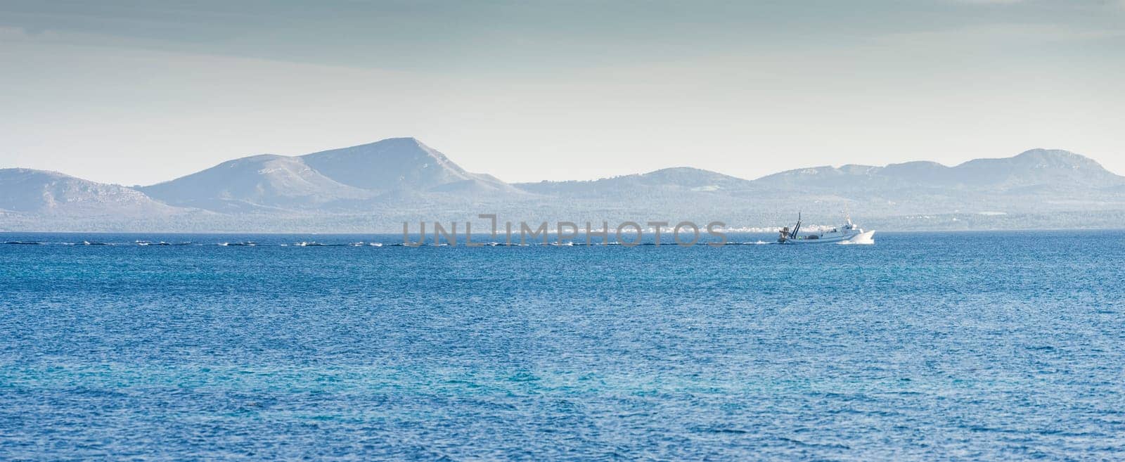 A fishing trawler cuts through the cobalt blue waters, its wake creating a frothy trail, set against the serene backdrop of distant mountains under a clear sky.