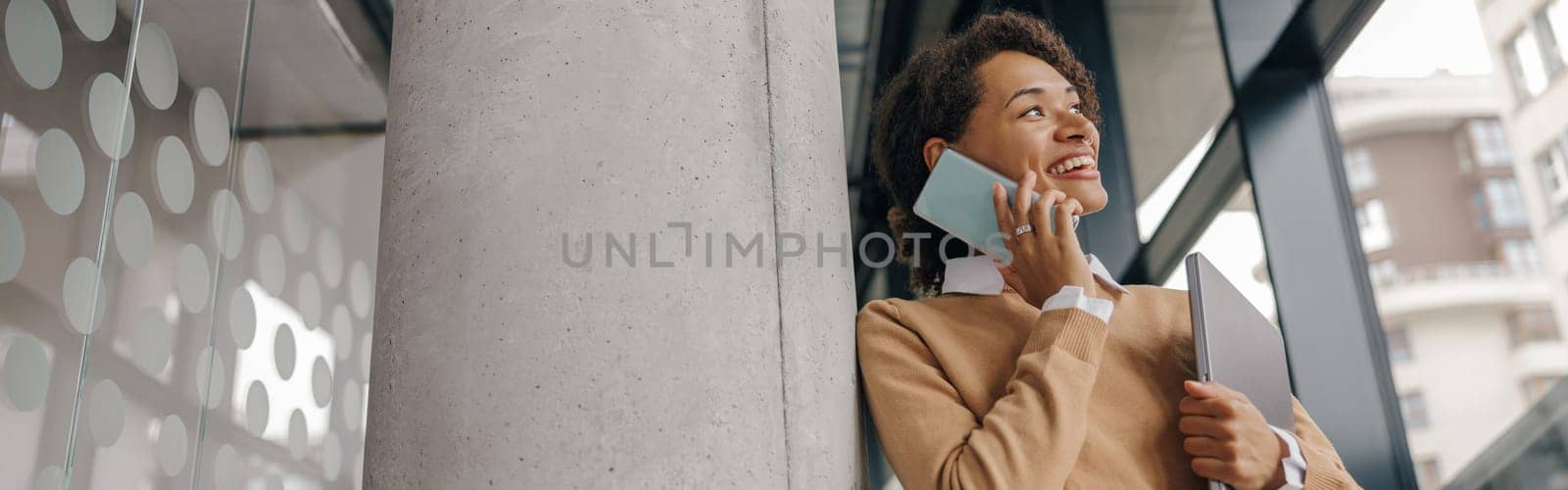 Female manager is talking mobile phone while standing with laptop in modern office hall near window by Yaroslav_astakhov