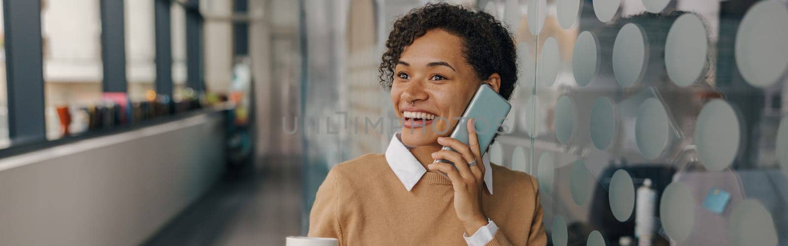 Smiling female manager with coffee cup is talking phone while standing on modern office background by Yaroslav_astakhov