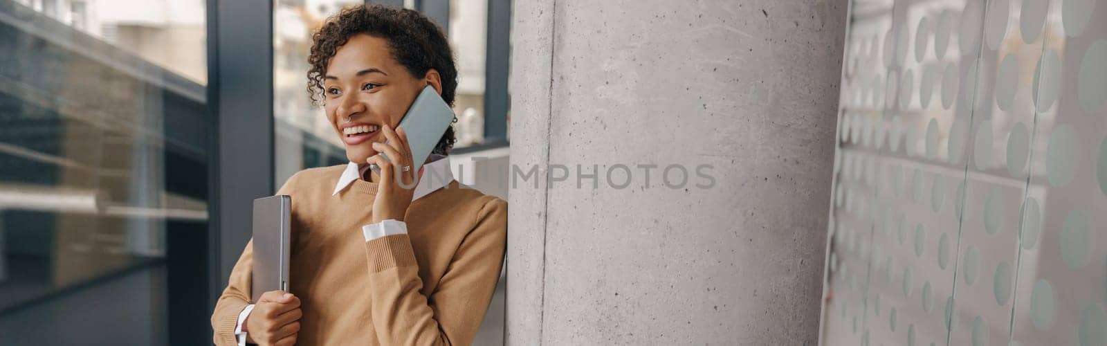 Stylish business woman is talking phone while standing with laptop in modern office