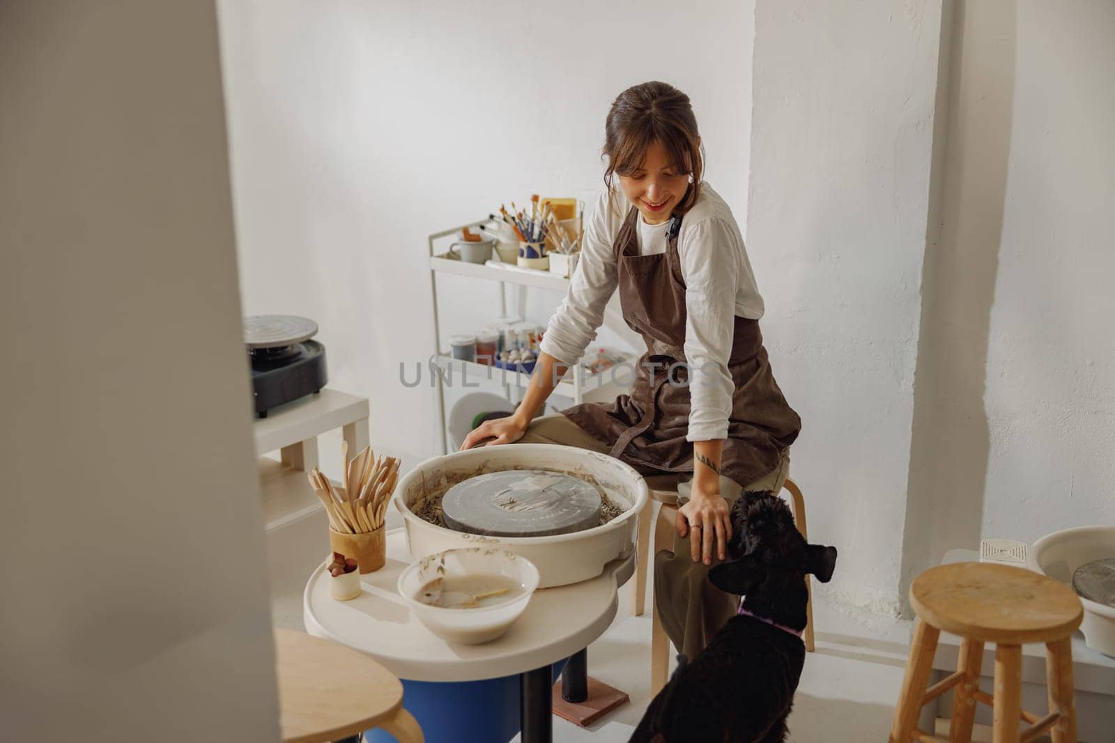 Smiling female ceramist wearing apron with her dog working in pottery studio by Yaroslav_astakhov