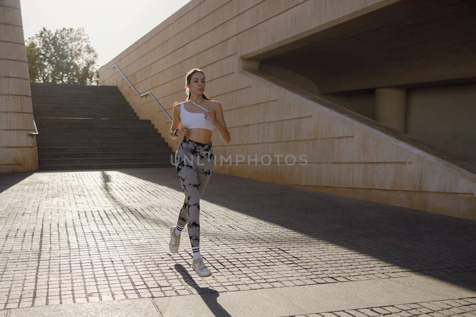 Young woman in sportswear is running on modern buildings background. Active lifestyle concept by Yaroslav_astakhov