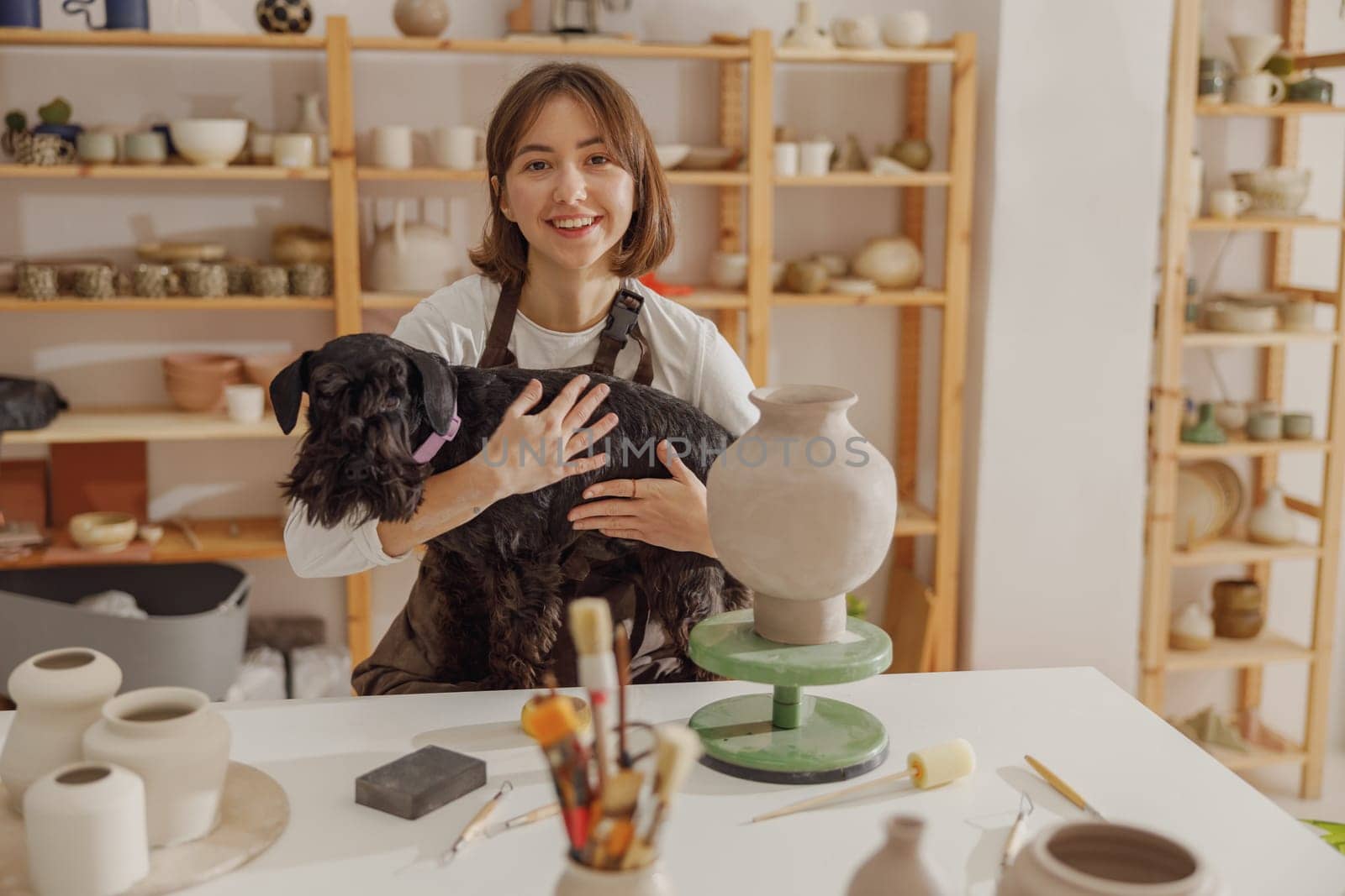 Smiling female ceramist holding her dog while sitting in pottery studio. High quality photo