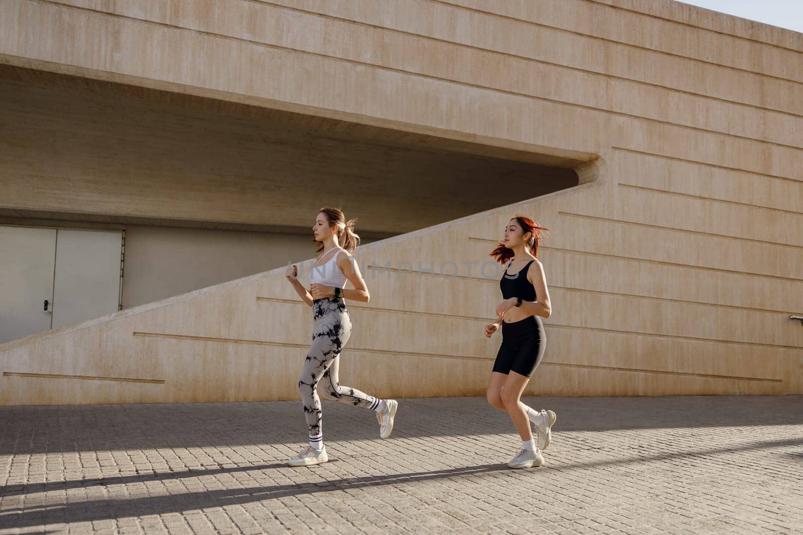 Two smiling women athlete running side by side along an outdoor track on modern buildings background by Yaroslav_astakhov