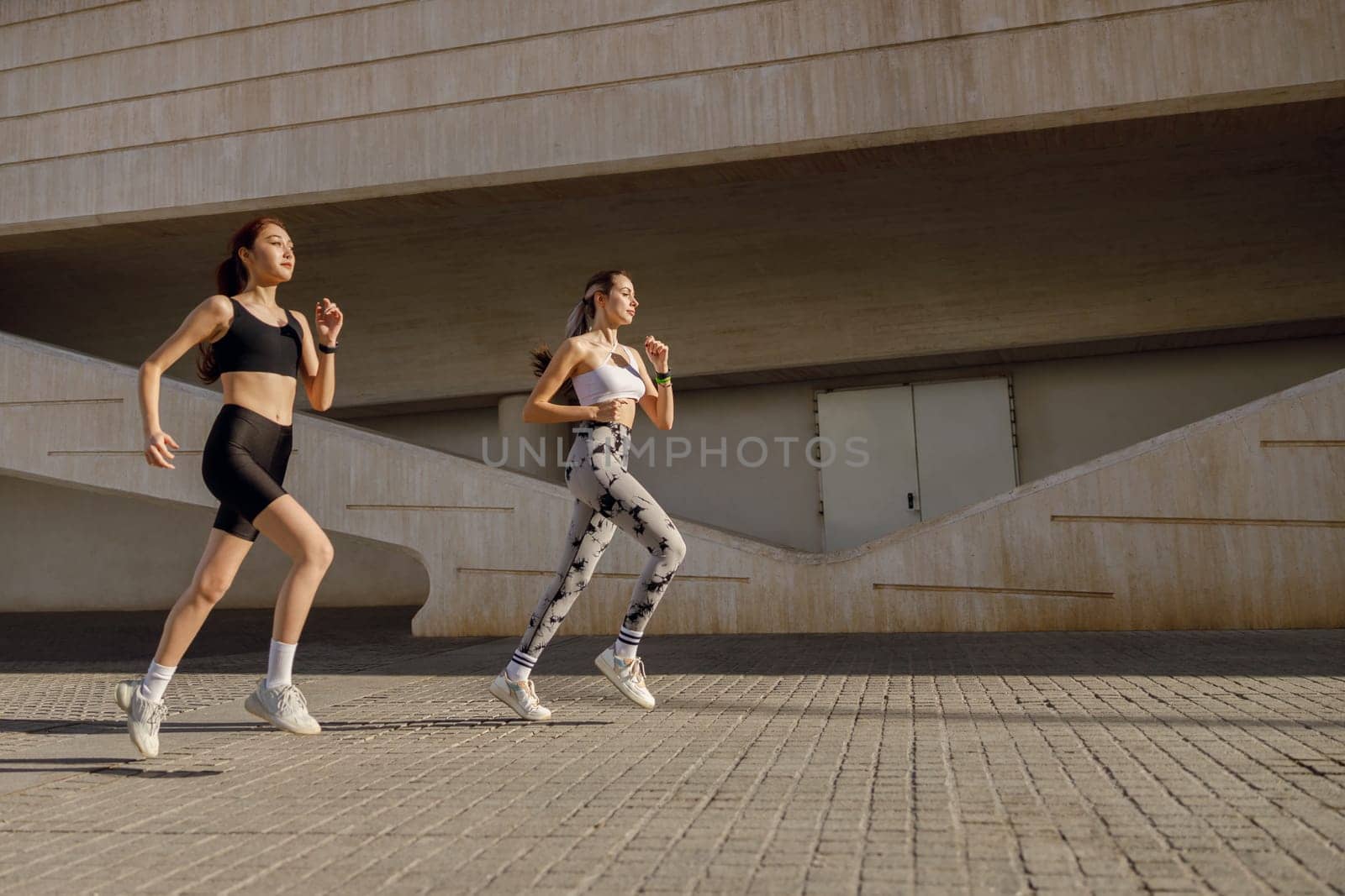 Two young women in sportswear are running on modern buildings background. Active lifestyle concept