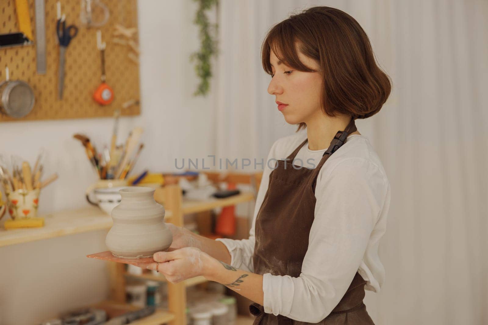 Professional potter wearing apron holding unfired clay vase while standing in pottery studio