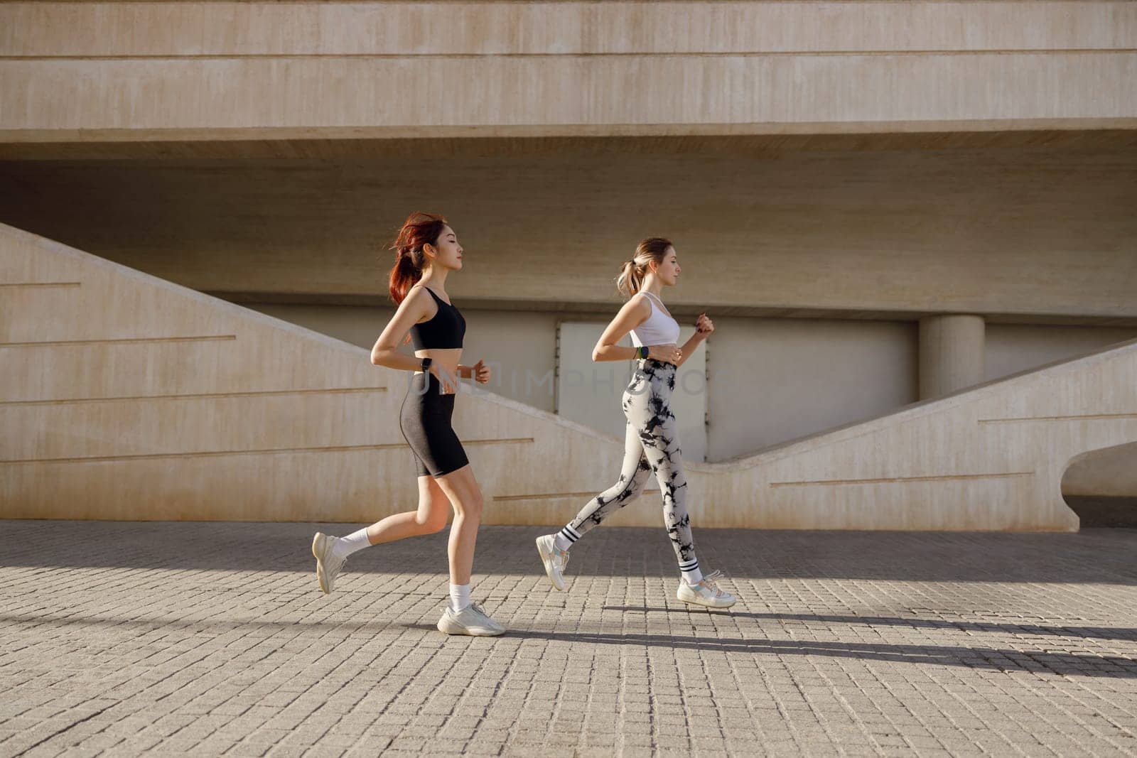 Two young women in sportswear are running on modern buildings background. Active lifestyle concept by Yaroslav_astakhov