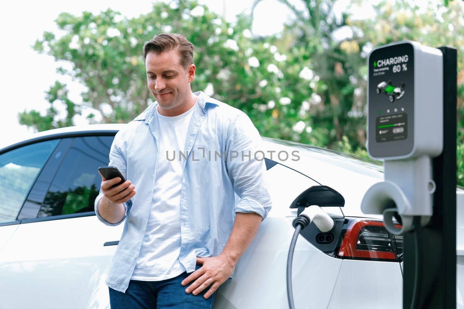 Modern eco-friendly man recharging electric vehicle from EV charging station, using Innovative EV technology utilization for tracking energy usage to optimize battery charging on smartphone.Synchronos