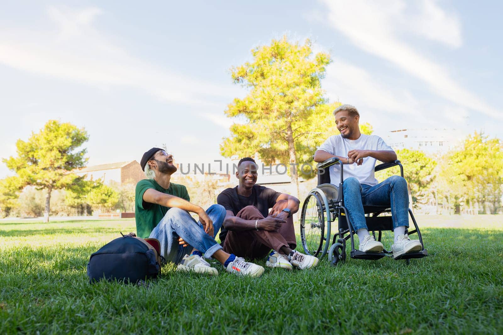 Young African American man in a wheelchair and his male friends laughing and having fun together sitting on the grass in a park. Copy space. Friendship and disability.