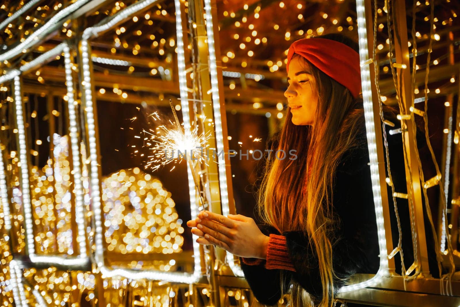 Woman holding sparkler night while celebrating Christmas outside. Dressed in a fur coat and a red headband. Blurred christmas decorations in the background. Selective focus.