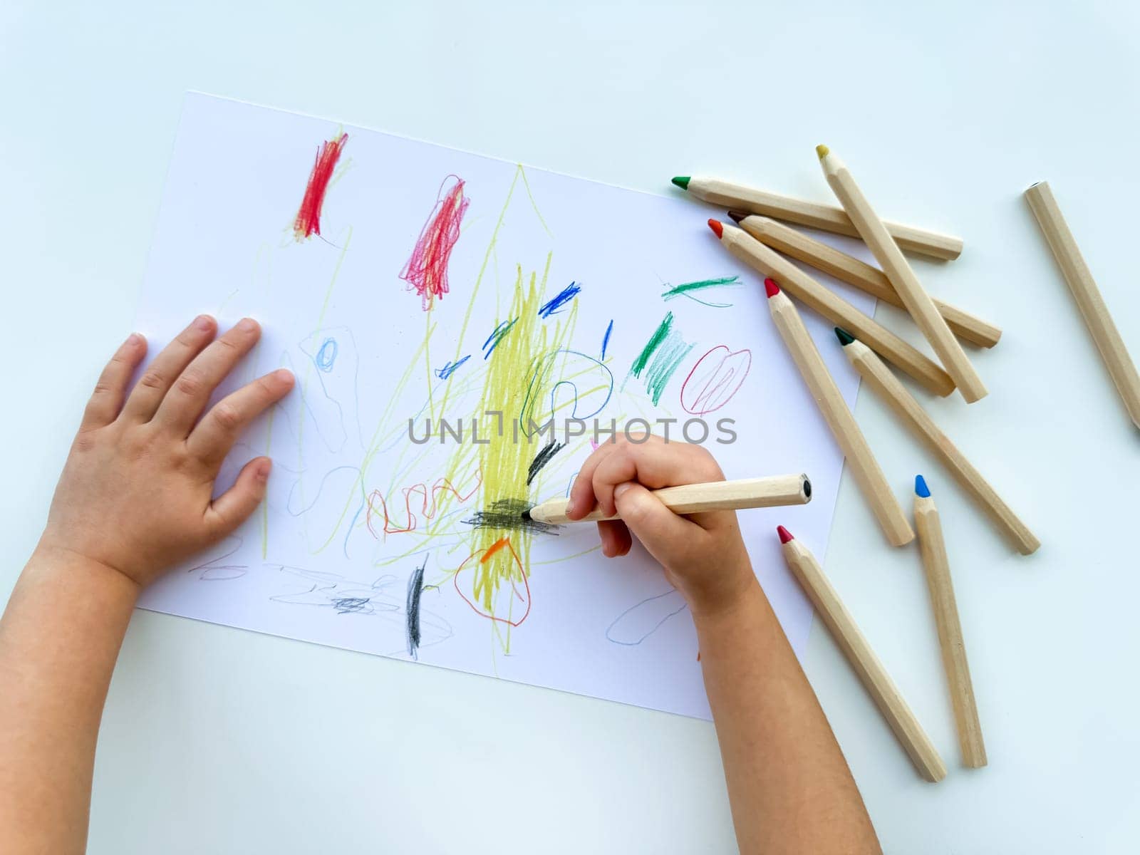 small child draws with colored pencils on paper on white table. High quality photo