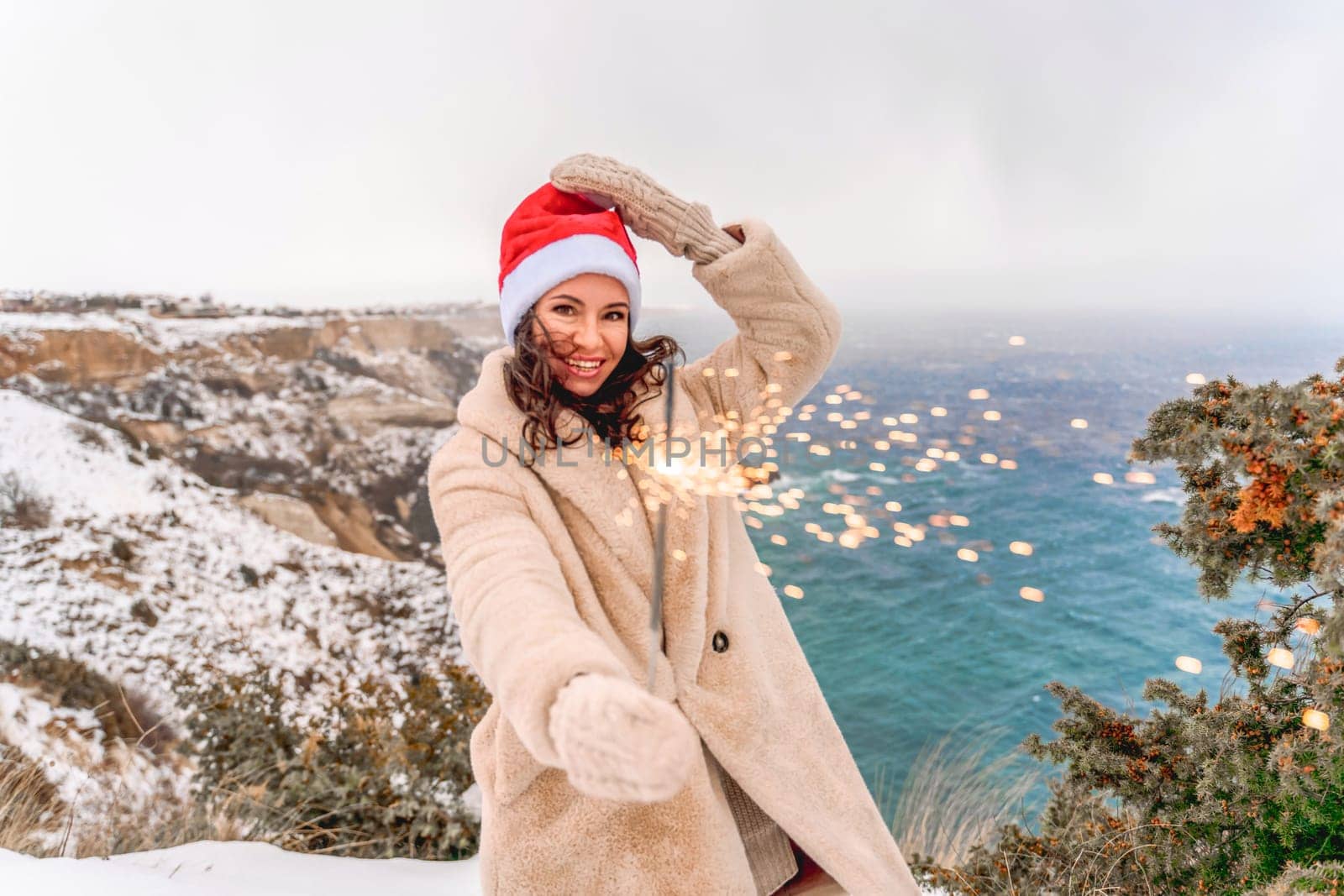 Outdoor winter portrait of happy smiling woman, light faux fur coat holding heart sparkler, posing against sea and snow background.