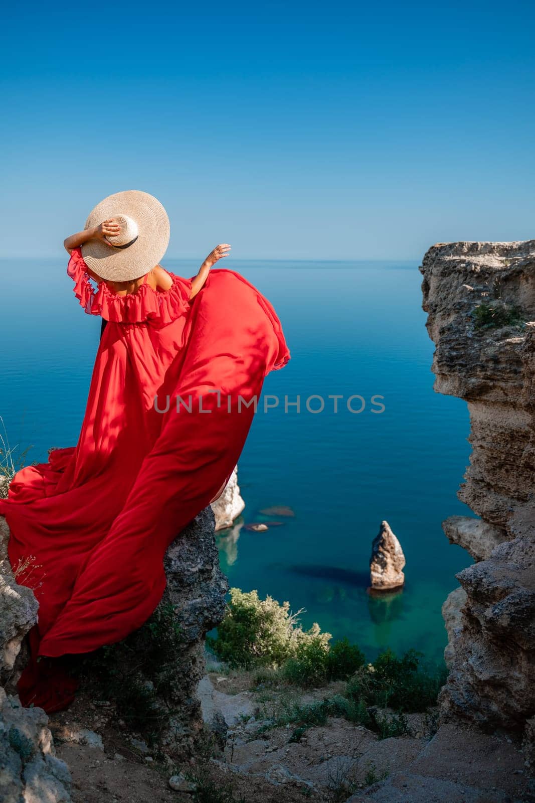Red dress sea woman. Happy woman with flowing hair in a long flowing red dress stands on a rock near the sea. Travel concept, photo session at sea by Matiunina
