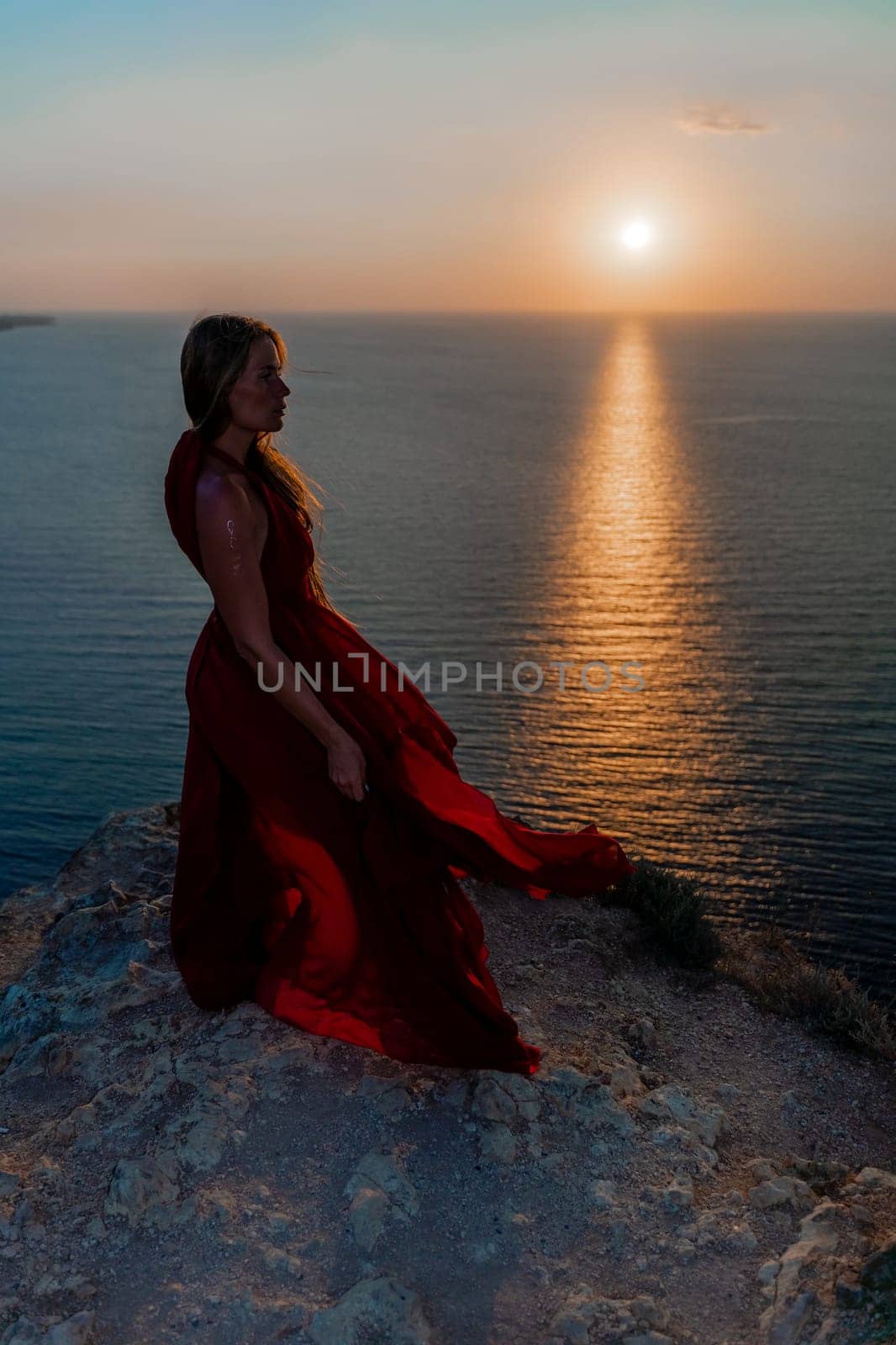 Woman sunset sea red dress, back view a happy beautiful sensual woman in a red long dress posing on a rock high above the sea on sunset