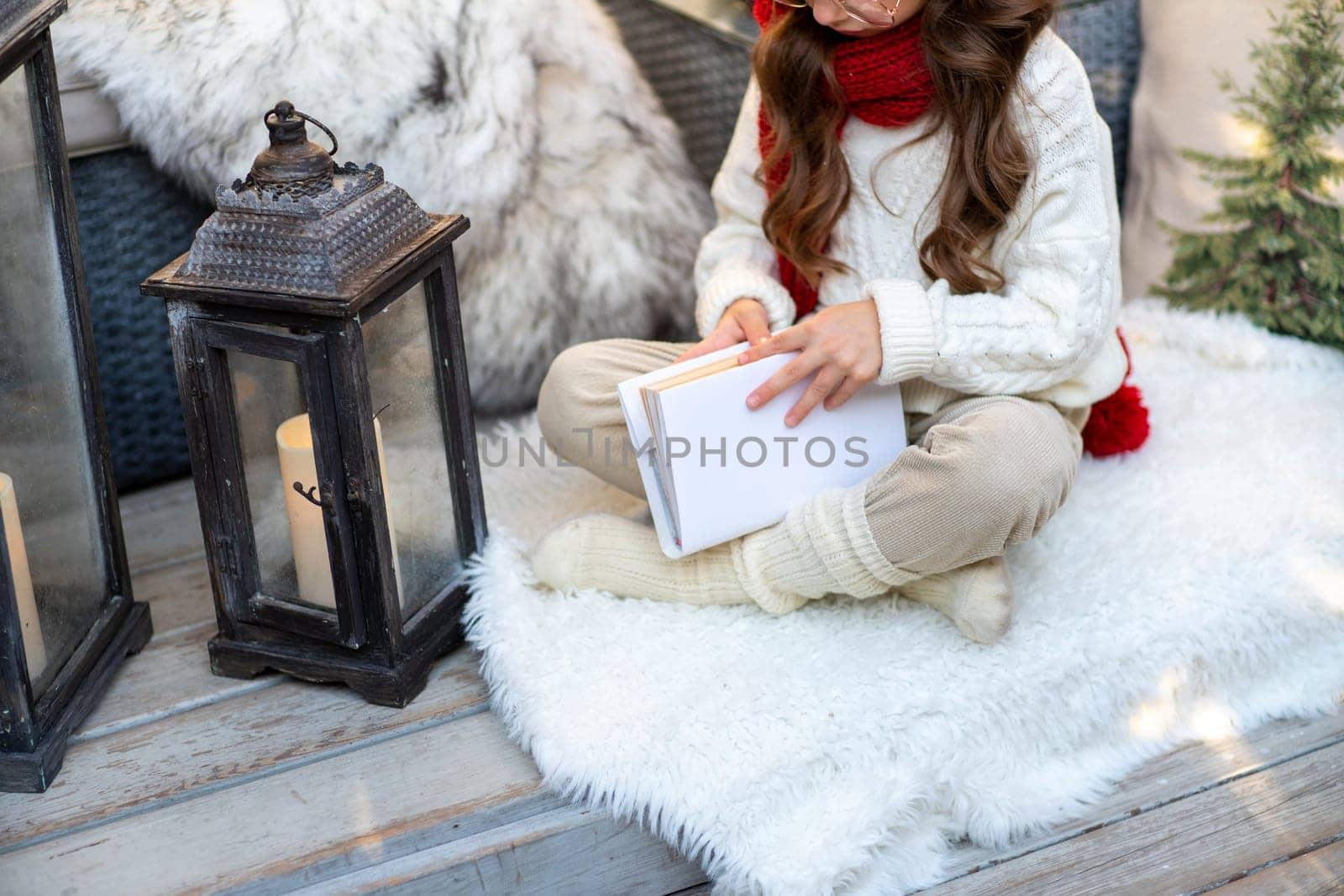 A girl with a book is sitting on the floor on a furry white carpet. Dressed in a white sweater and light trousers with a red scarf