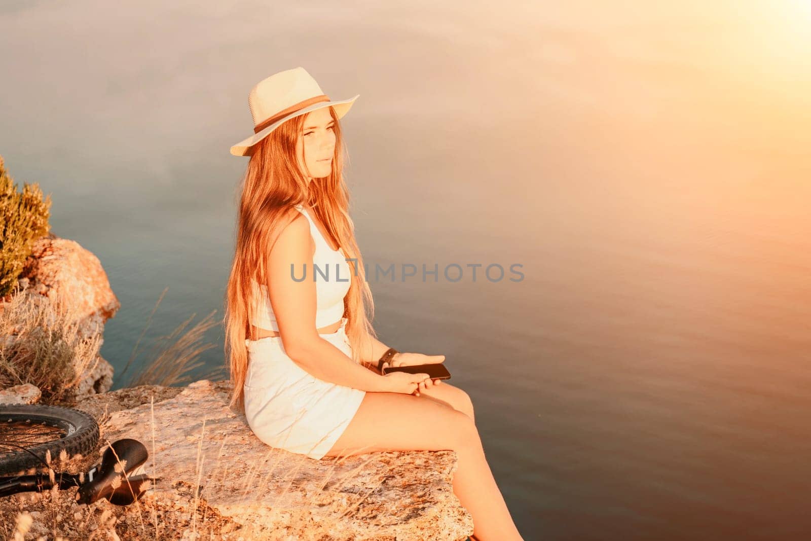 A tourist woman is sitting by the sea in a hat and white summer clothes, looking happy and relaxed