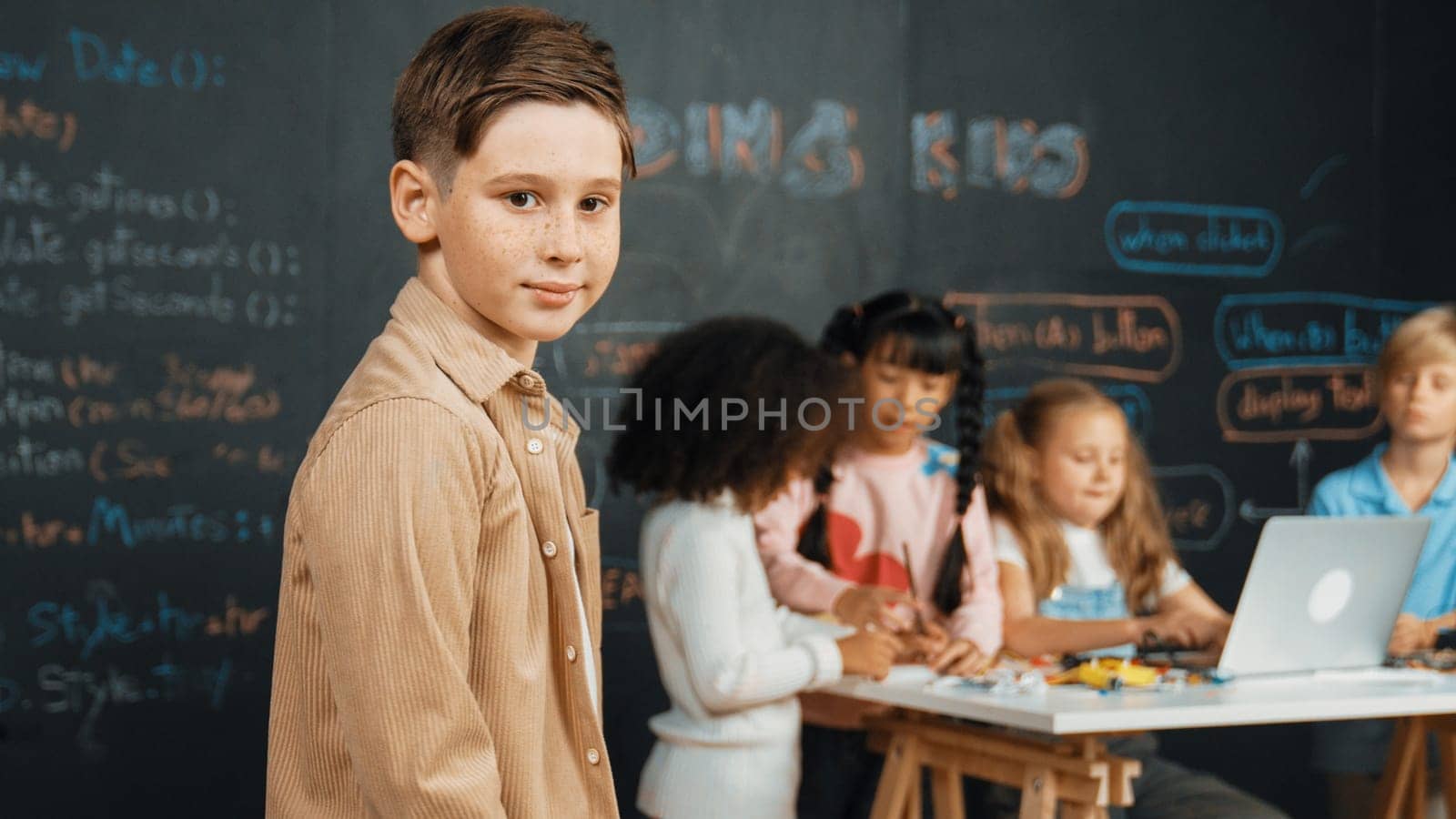 Smart boy smiling at camera while diverse friend working or learning engineering code or prompt in STEM technology classroom. Cute student standing at camera while children using laptop. Erudition.