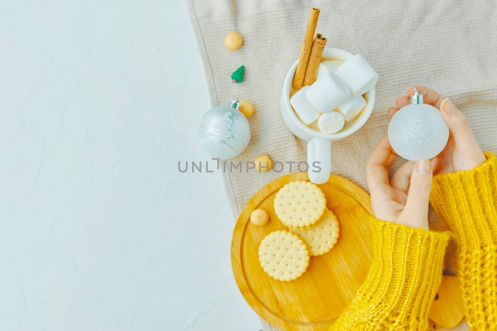 Woman in a yellow sweater drinks hot cocoa with marshmallow and cinnamon rolls in a white mug and cookies and a figure of a Christmas tree