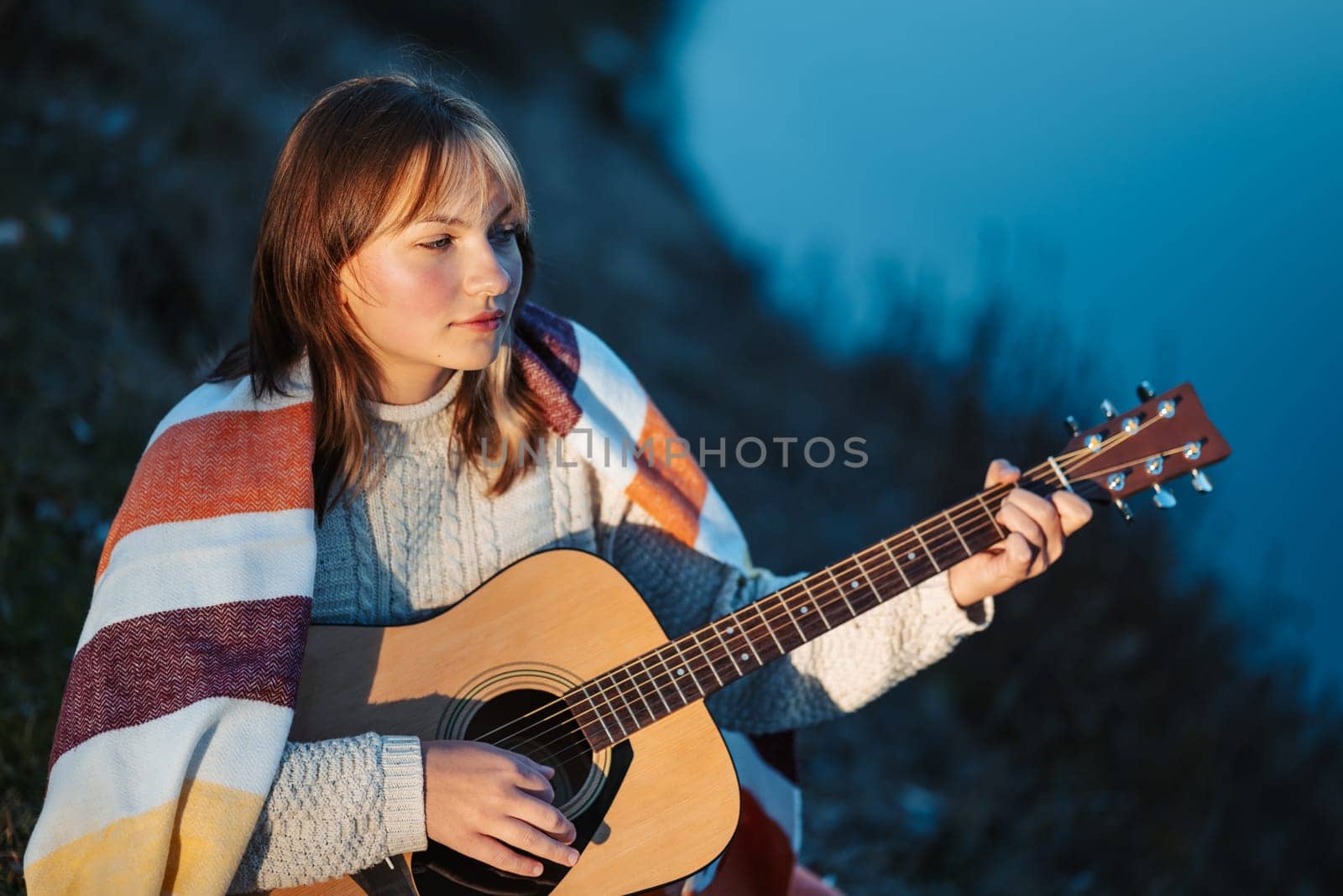 Girl sitting on river bank and practising playing the guitar, mastering skills, learning new composition