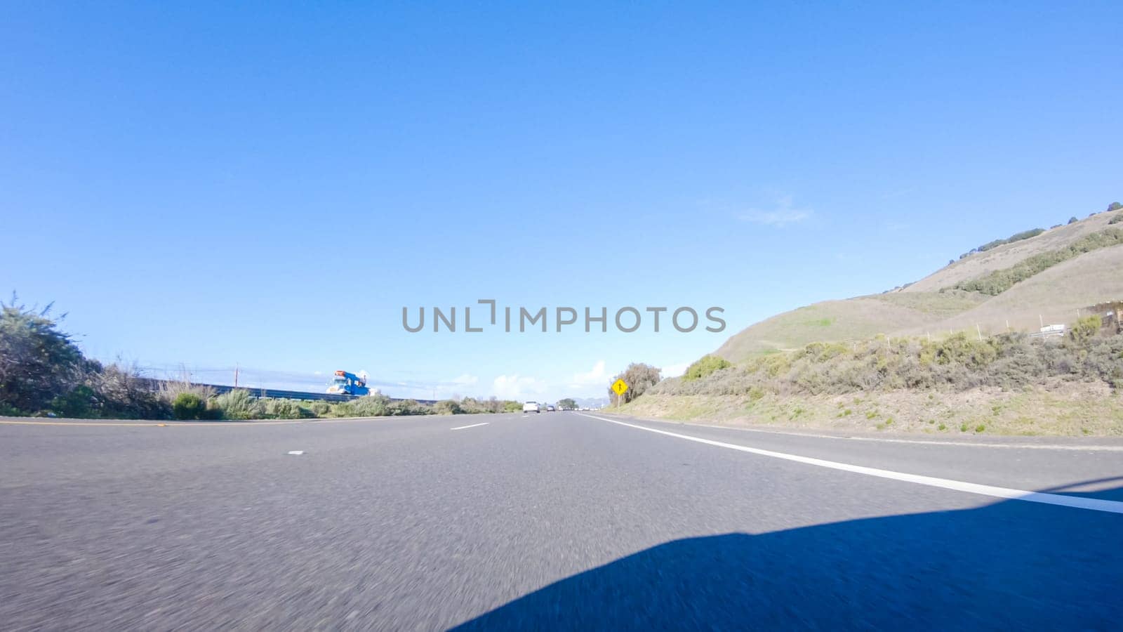 On a crisp winter day, a car cruises along the iconic Highway 1 near San Luis Obispo, California. The surrounding landscape is brownish and subdued, with rolling hills and patches of coastal vegetation flanking the winding road.