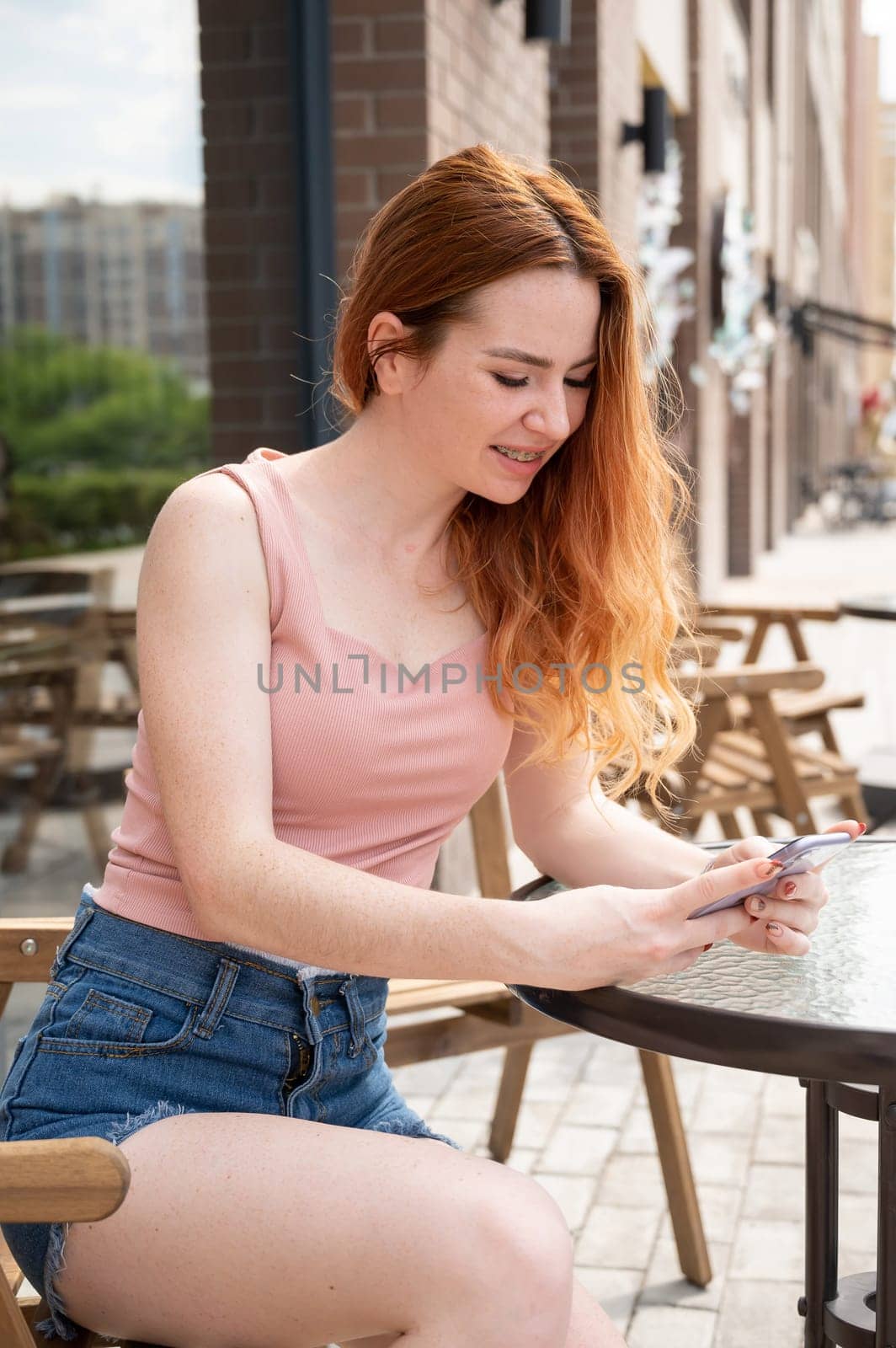 Young woman with braces on her teeth smiles while sitting in an outdoor cafe and uses a smartphone. Vertical photo by mrwed54