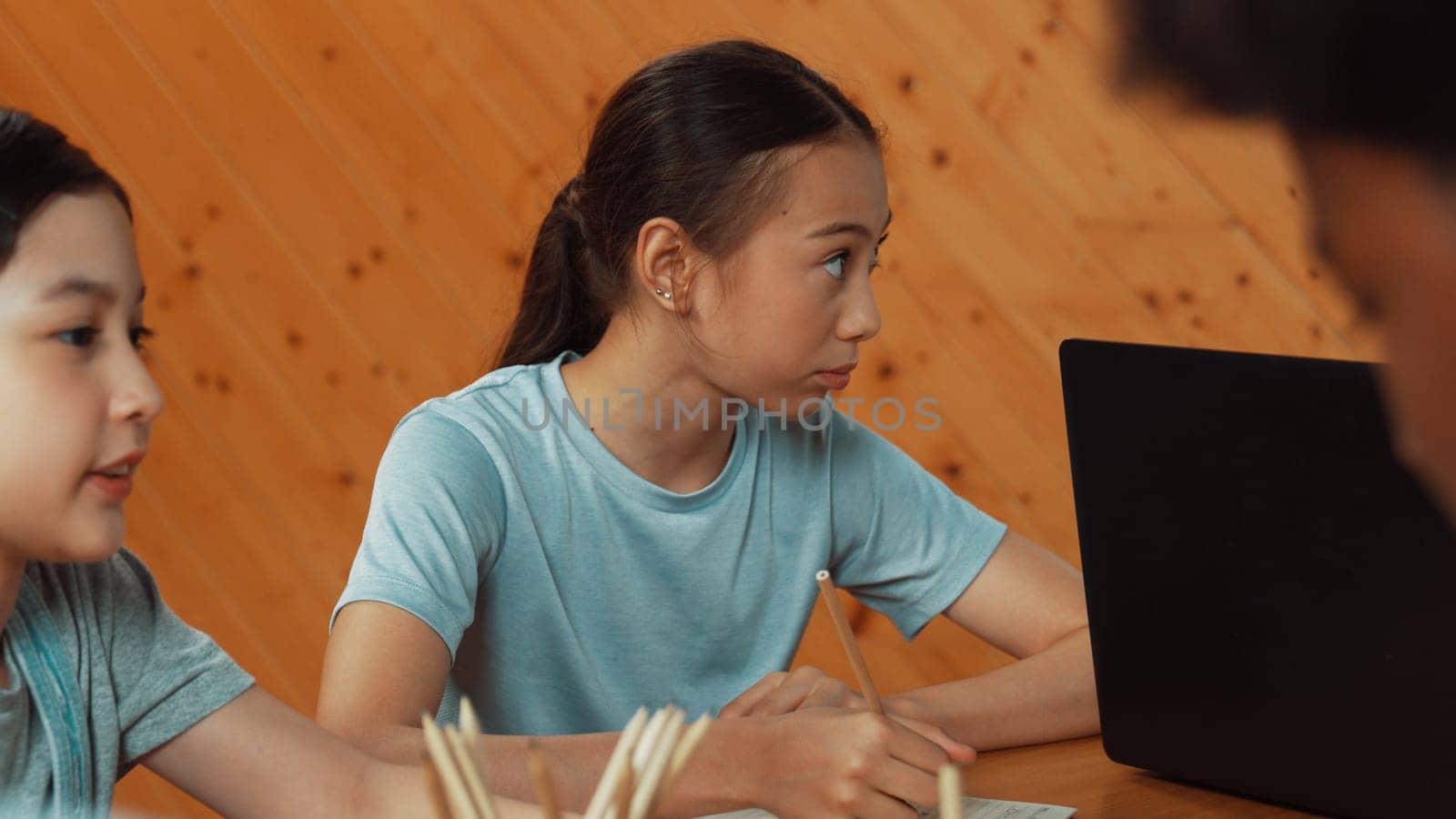 Closeup of happy asian cute girl writing answer down in paper or taking a note in classroom while diverse student working together on table with laptop placed. Creative education concept. Edification.