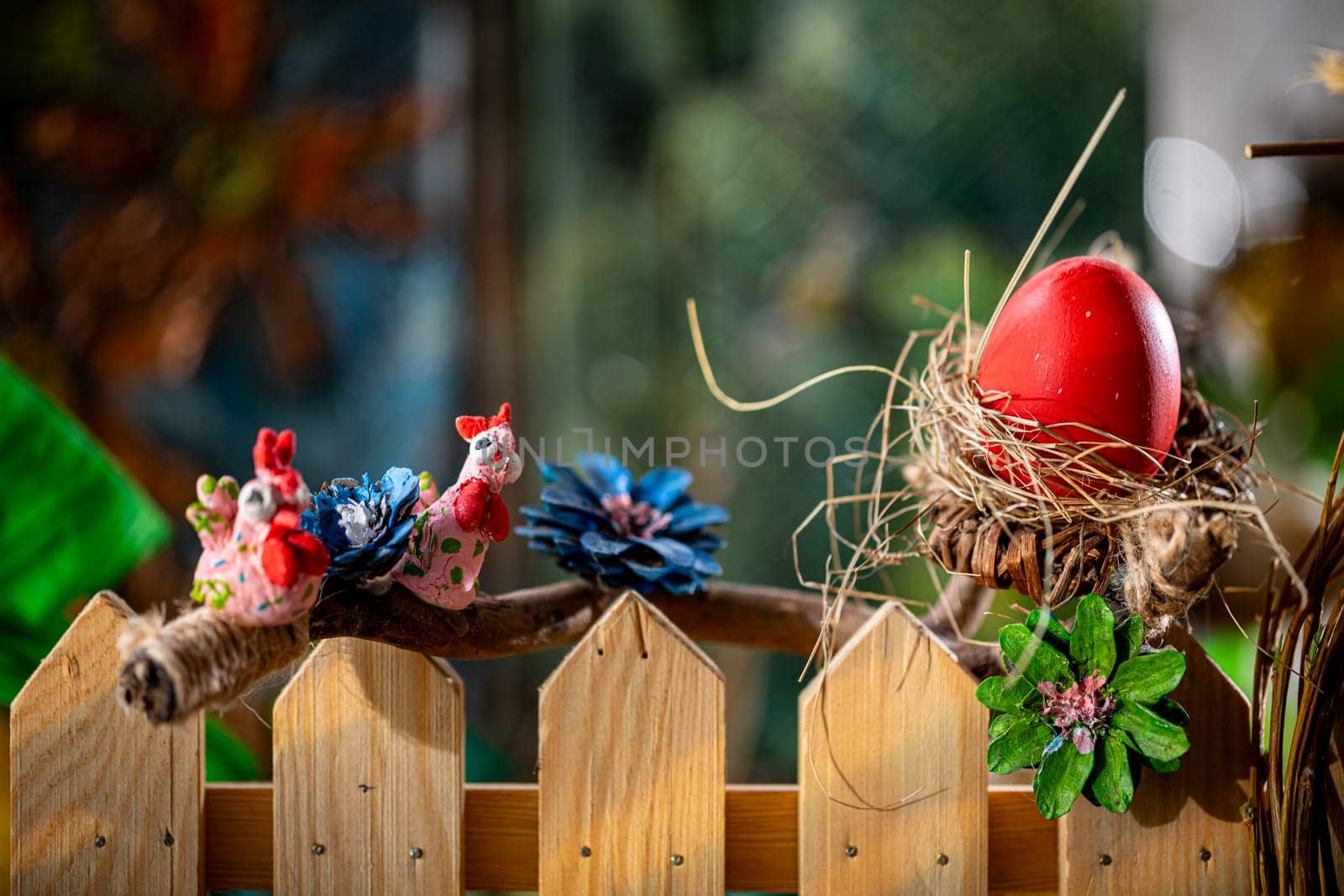 Amidst a wooden fence and arranged branches lies a nest intricately woven from dry straw, cradling a large egg painted in vibrant red.