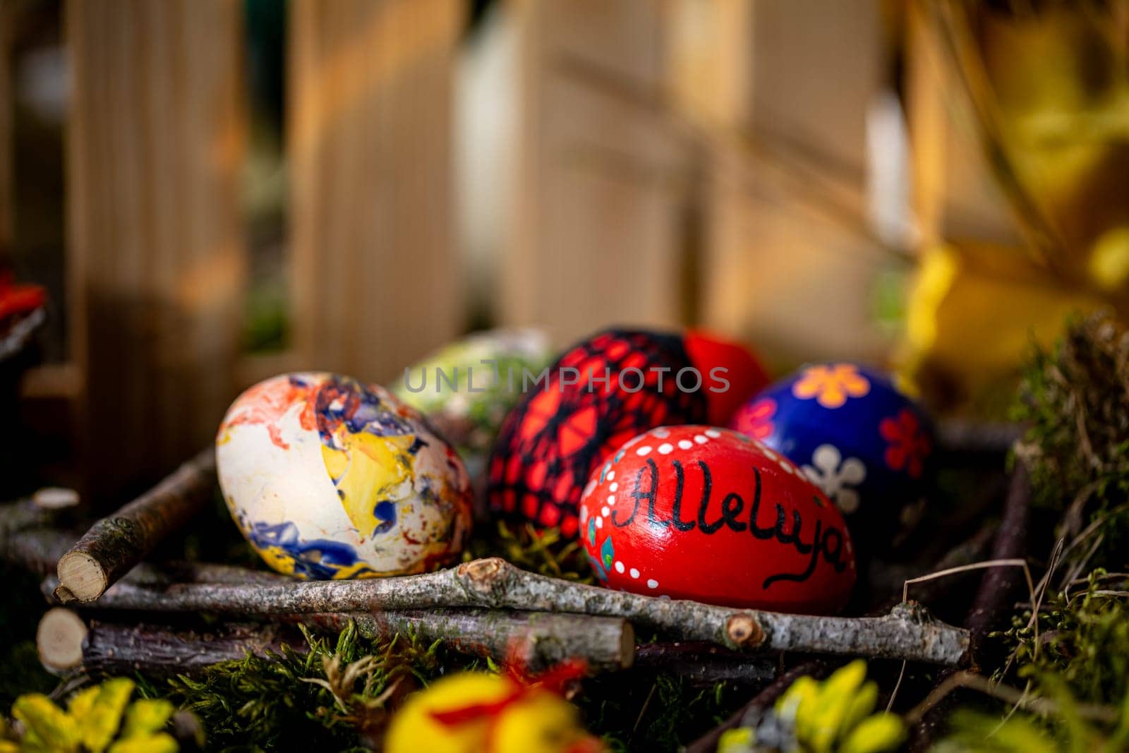 In this tranquil Easter scene, delicate handcrafted eggs rest in a basket lined with arranged twigs and lush green moss. One egg bears the inscription ,Alleluja, while the others feature floral patterns or colorful artistic doodles.