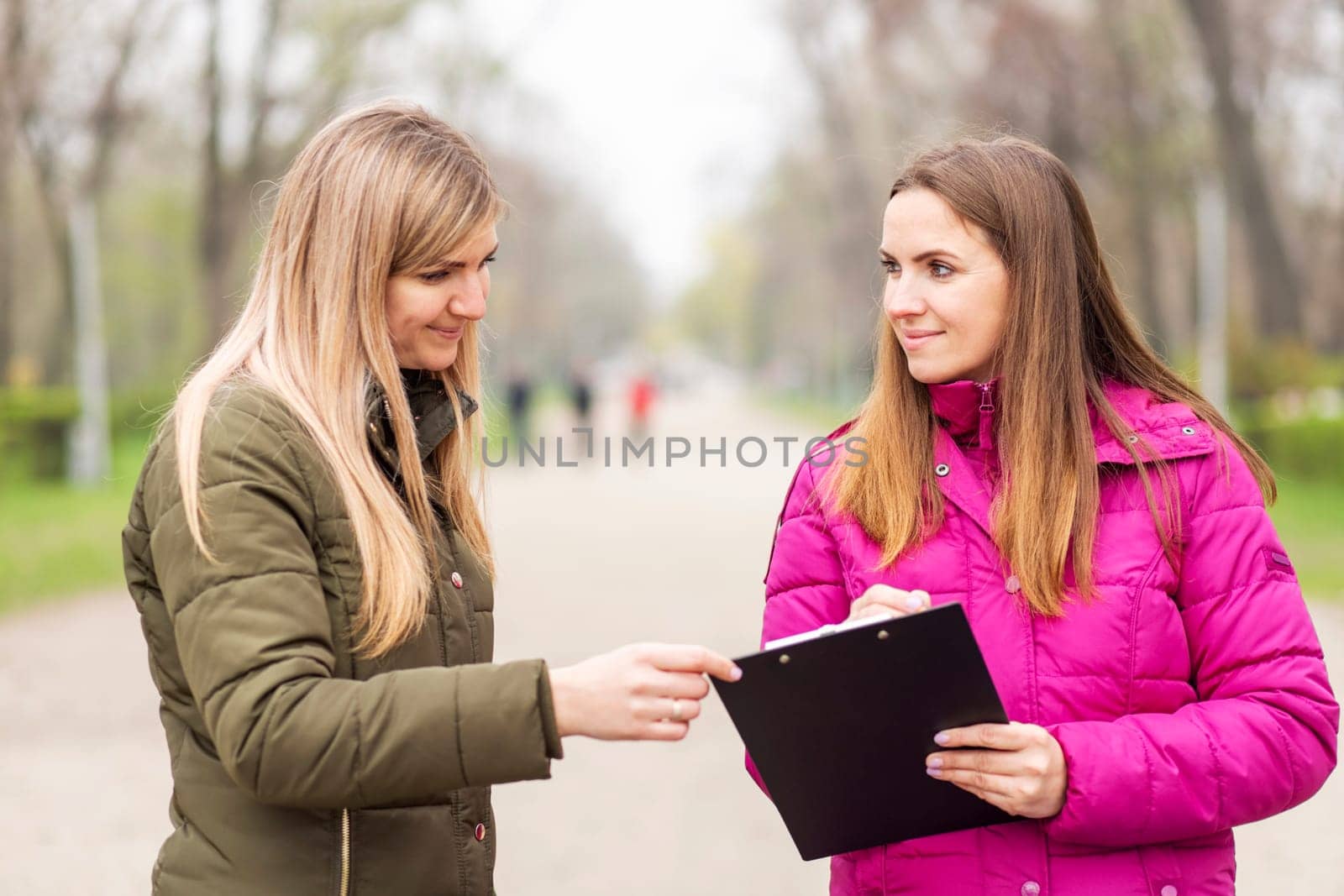 Two women engaged in a conversation with a clipboard on a park path.