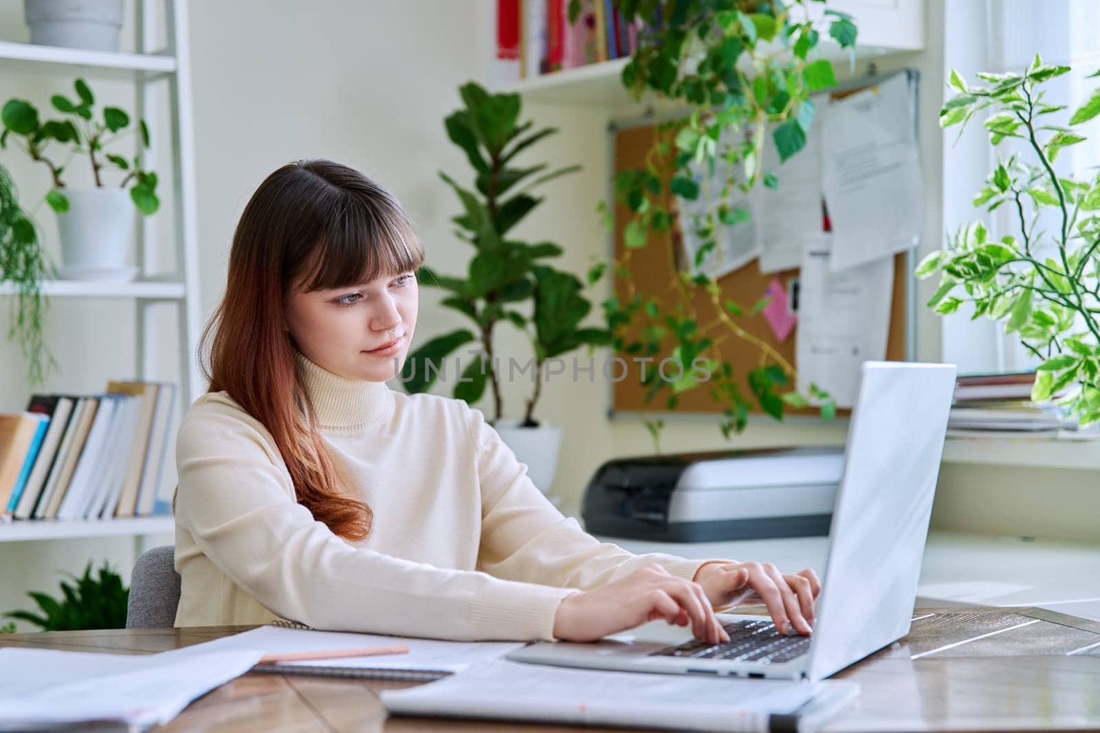 Young female college student studying at home at desk using computer laptop, typing on a keyboard. E-learning, education, technology, knowledge, youth concept